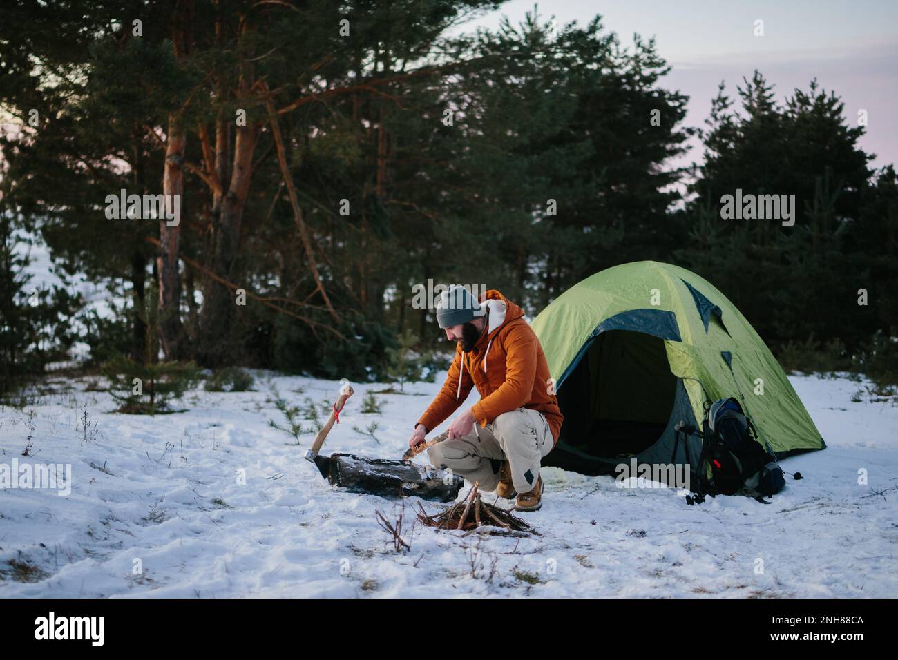 Un uomo seduto vicino alla sua tenda prepara lana di legno con un coltello per avviare un fuoco in una foresta invernale al tramonto. Concetto di sopravvivenza invernale. Foto Stock