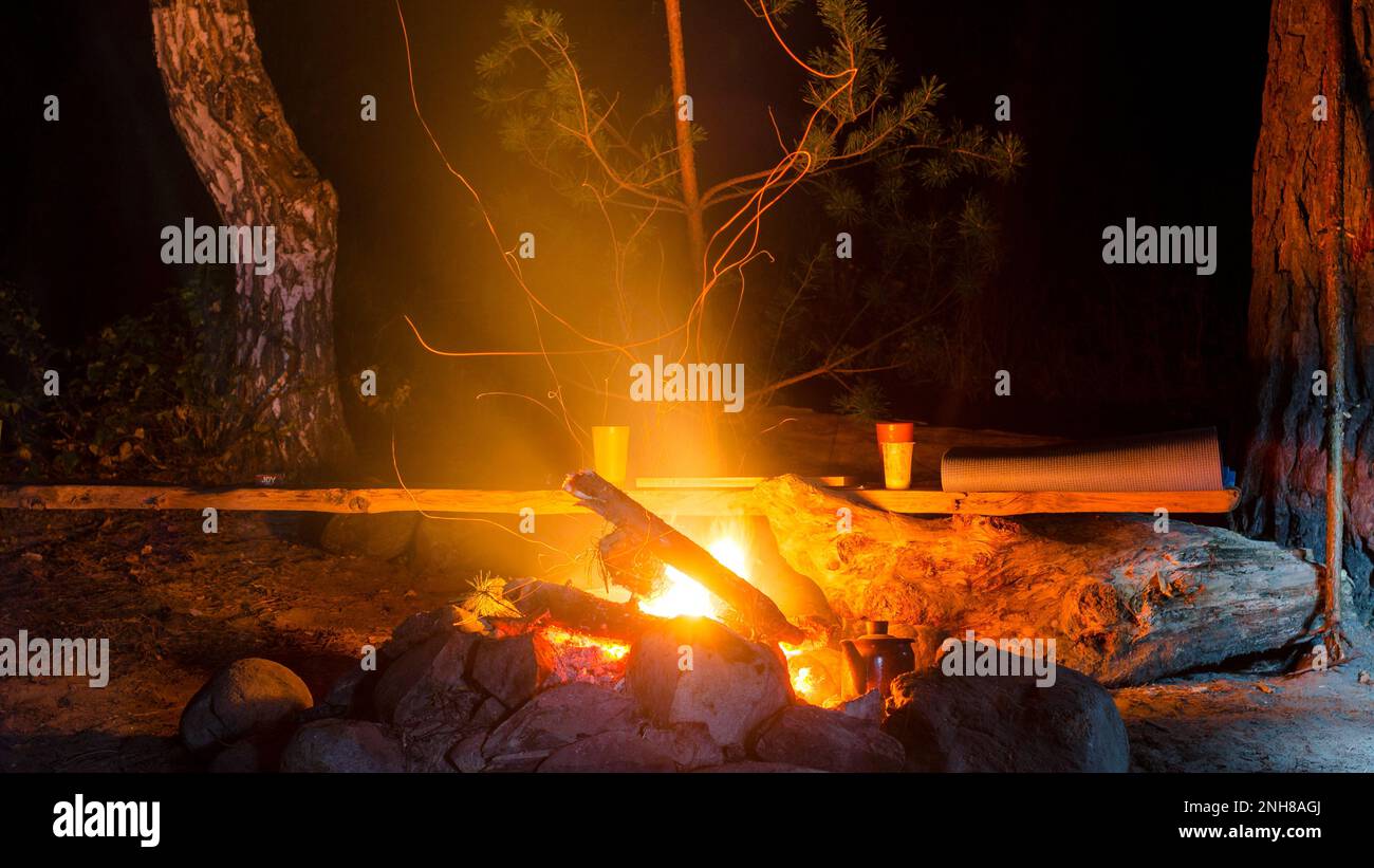 Coppe, backgammon e tappetino riposante si posano sulla panchina dal Consiglio di Amministrazione al Parco Turistico di notte vicino ad un fuoco che brucia nelle pietre con scintille Foto Stock