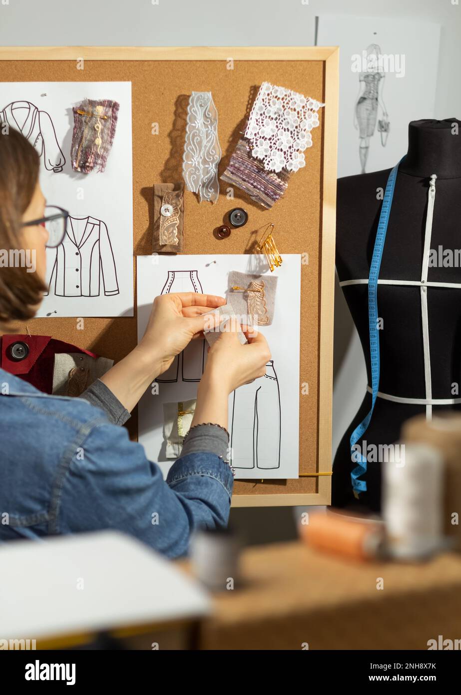 Una donna designer lavora con schizzi e campioni di tessuto per cucire abiti su un pannello di sughero. Foto Stock