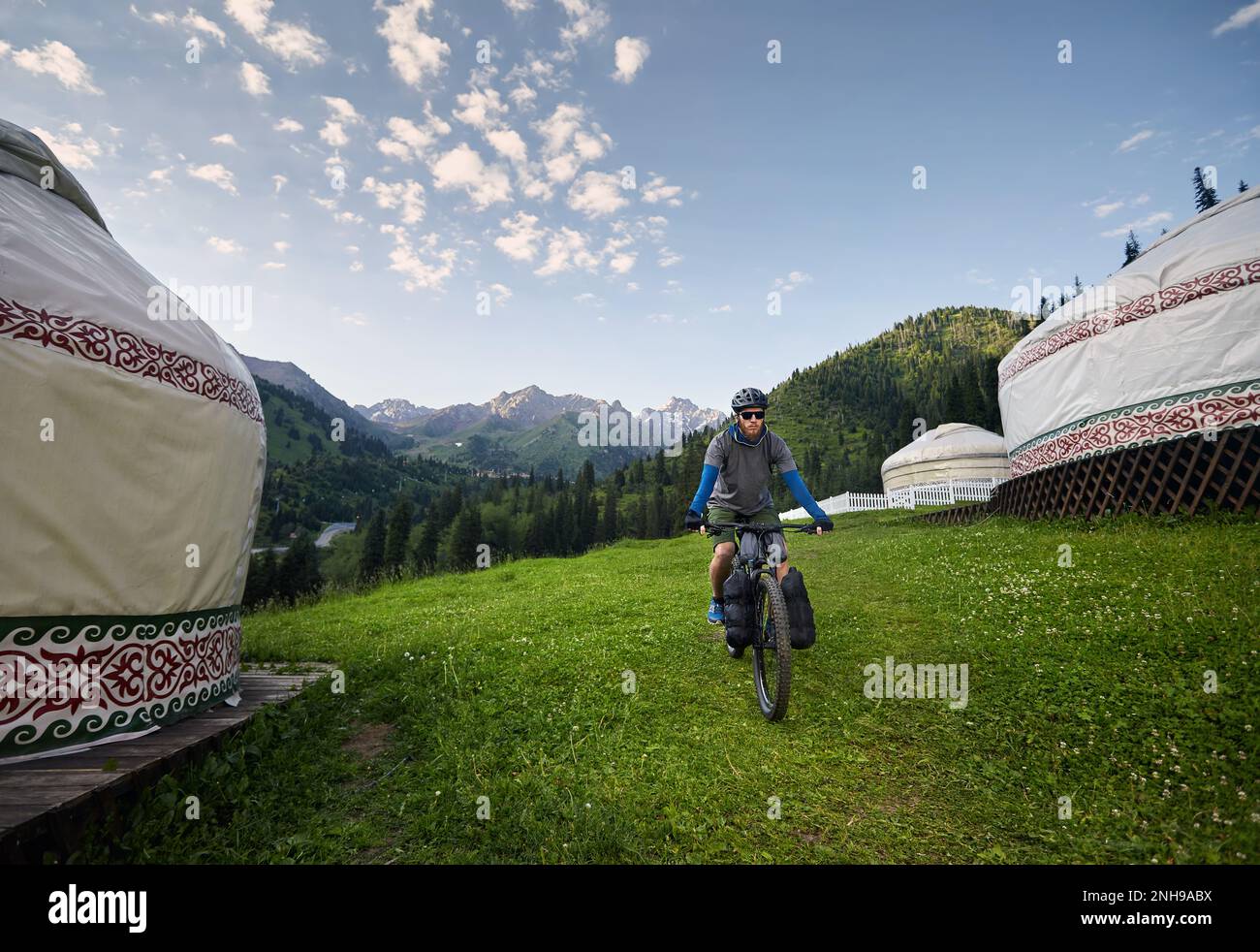 Uomo con greggi e barba in mountain bike con borse turistiche vicino a Nomad Yurt casa nella bella valle di montagna in Almaty, Kazakistan. Estrem Foto Stock