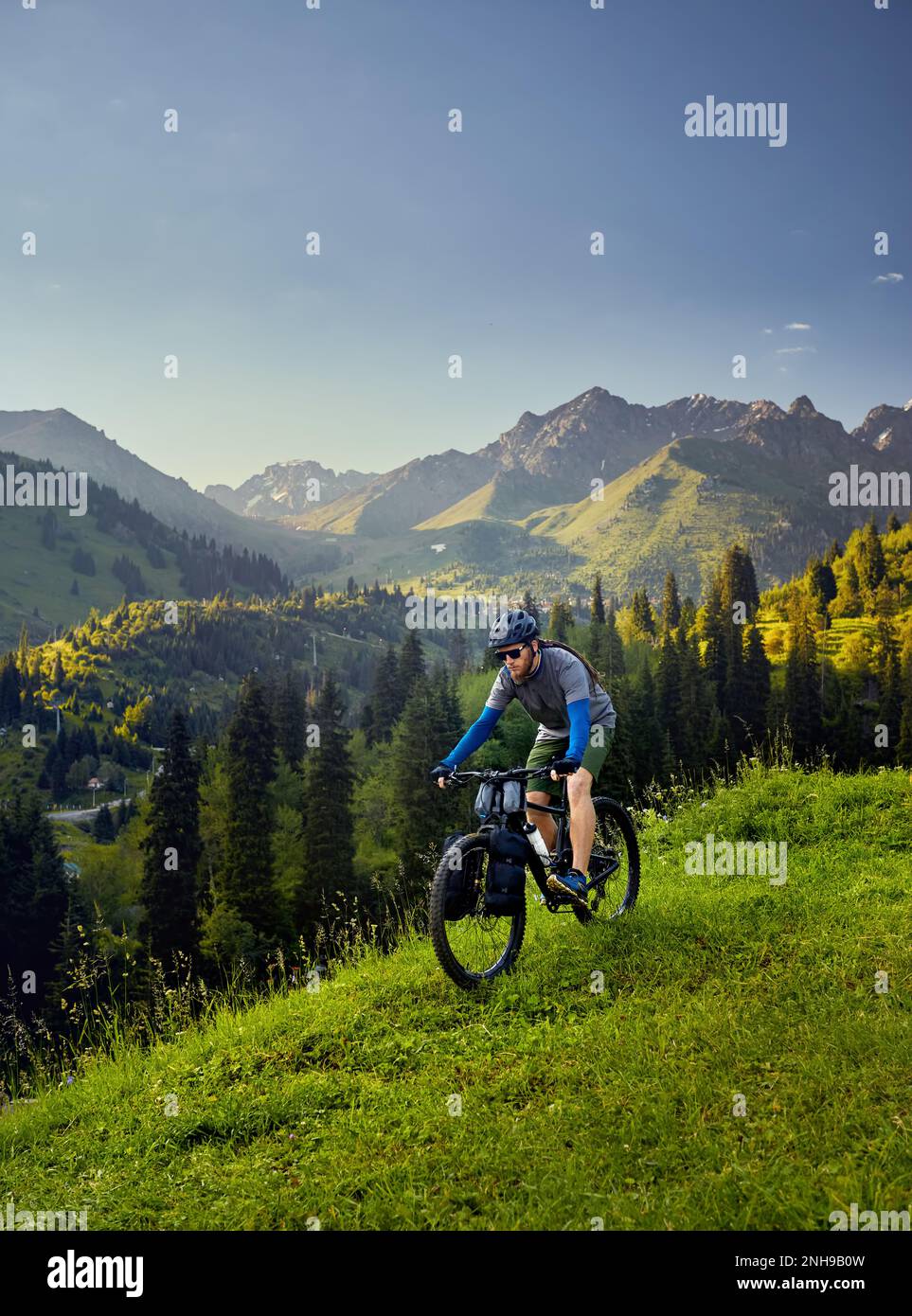 Uomo con greggi e barba giro in mountain bike con borse turistiche al verde foresta collinare nella bella valle di montagna al tramonto in Almaty, Kazakhistan Foto Stock