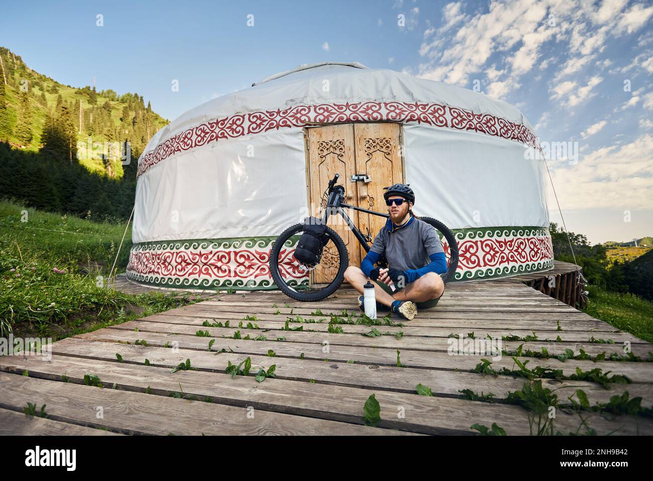 Uomo con greggi e barba seduto di fronte alla mountain bike con borse turistiche vicino a casa Nomad Yurt nella bella valle di montagna in Almaty, kazako Foto Stock