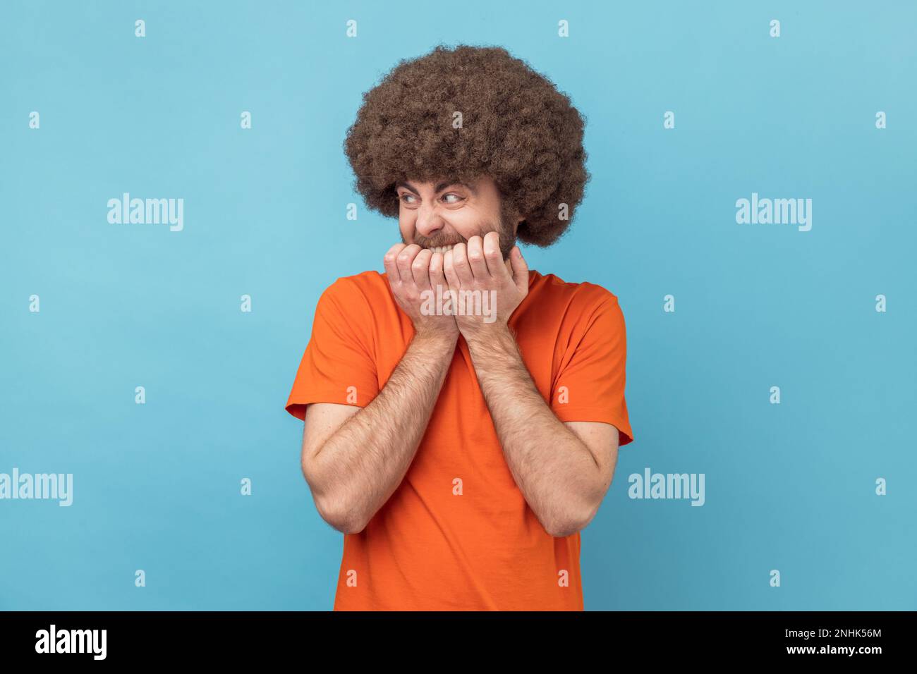 Disturbo d'ansia. Ritratto dell'uomo con acconciatura afro che indossa una T-shirt arancione mordente le unghie, nervosa per i disturbi, paurosa e spaventata. Studio in interni isolato su sfondo blu. Foto Stock