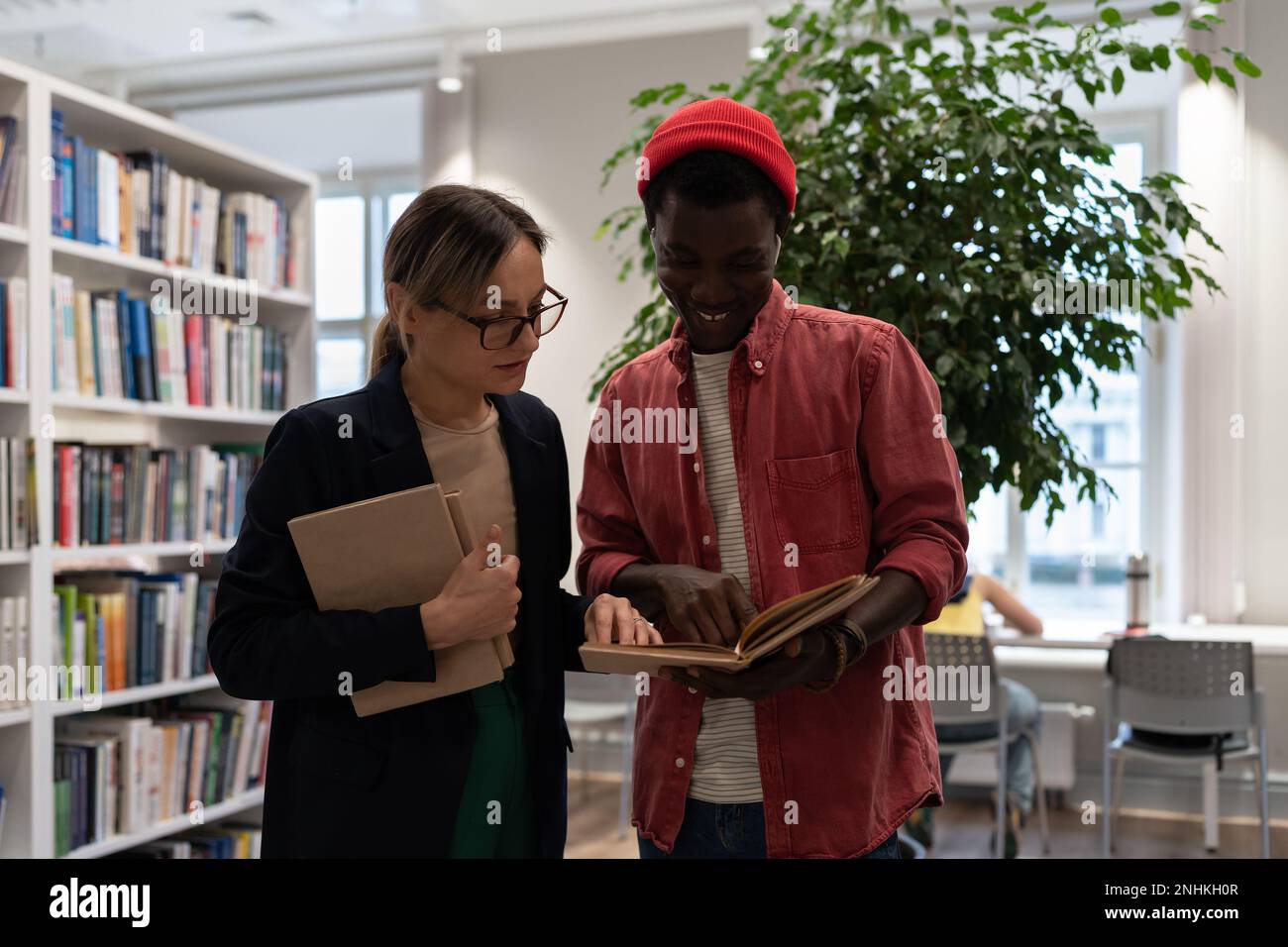 Professoressa donna concentrata che aiuta a capire il compito libro di testo per studente afroamericano uomo in biblioteca. Foto Stock