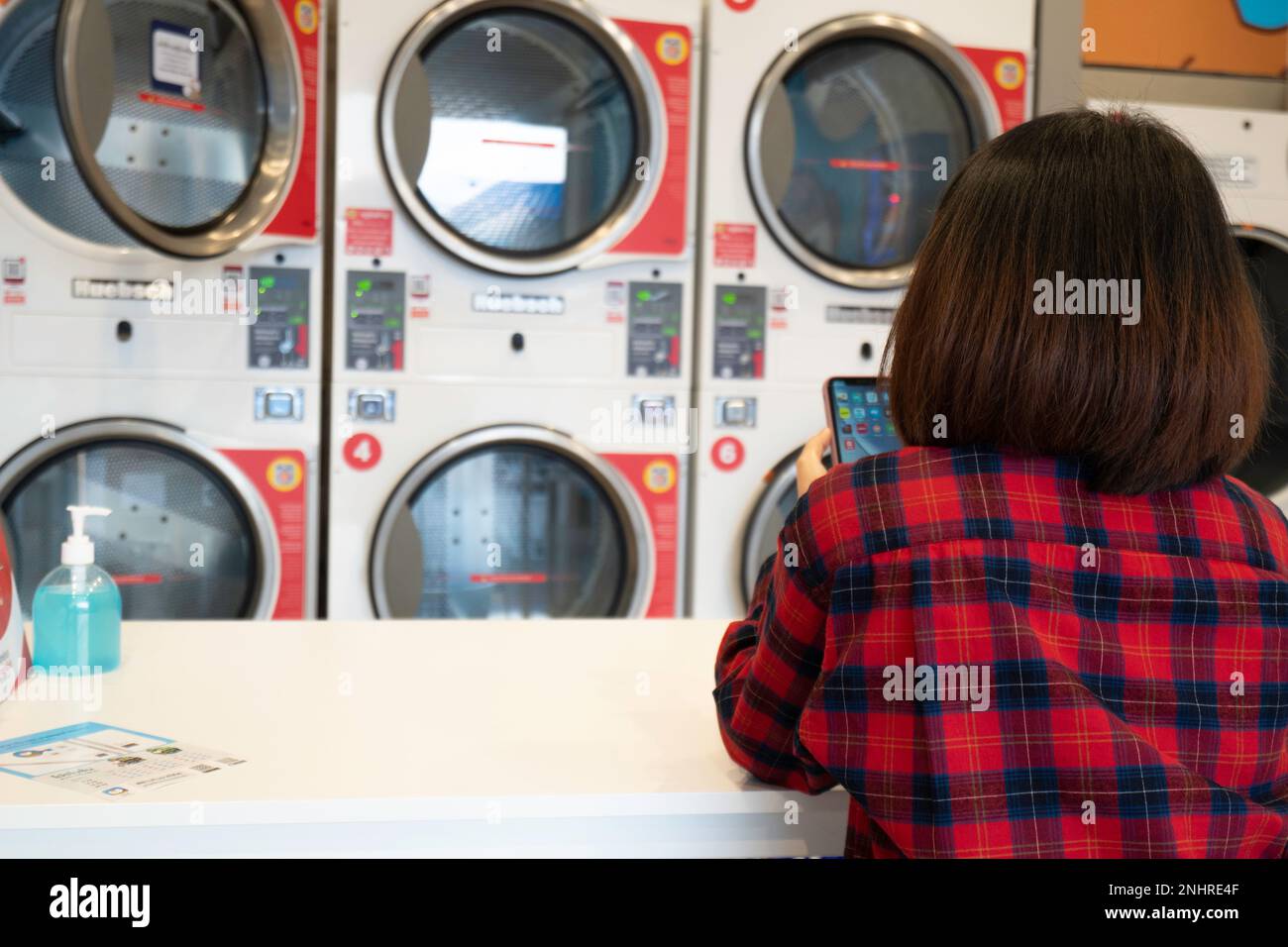 Vista posteriore della donna seduta con telefono cellulare in attesa di lavanderia lavata in lavatrice automatica presso il negozio di lavanderia automatica. Fila di lavaggio industriale mac Foto Stock
