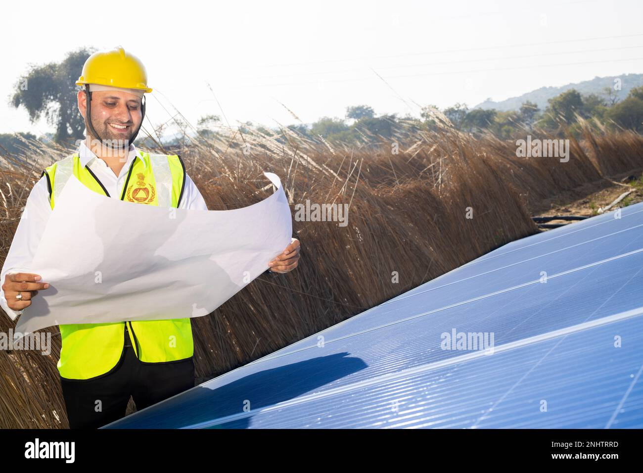 Giovane uomo indiano tecnico che indossa cappello giallo in piedi con carta da grafico pianificazione per installare solare panels.Industrial lavoratore impianto solare Foto Stock