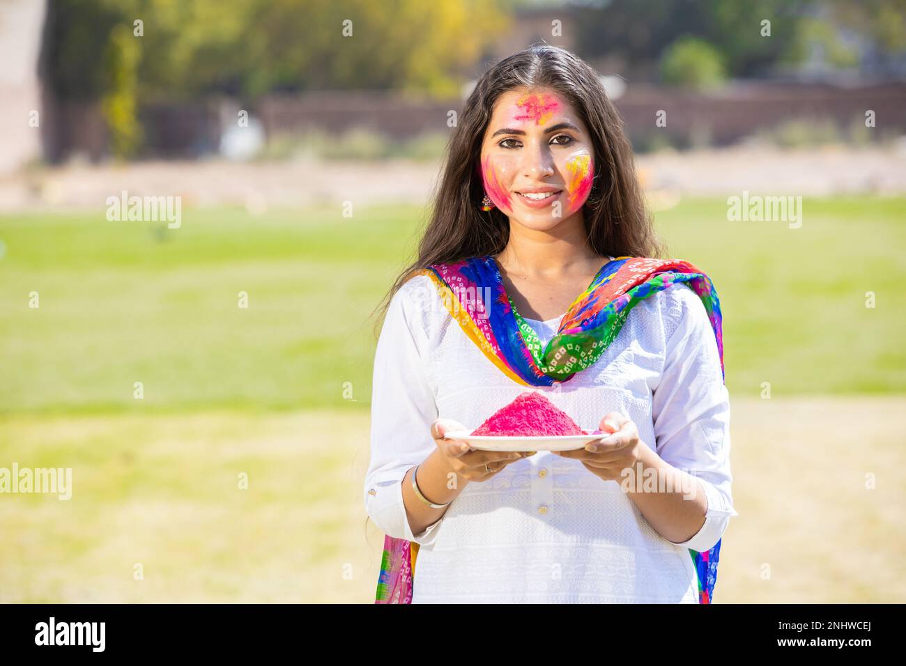 Ritratto di felice giovane donna indiana indossando kurta bianca e dupatta vestito con polvere di colore piatto che celebra il festival holi al parco all'aperto, faccia Foto Stock