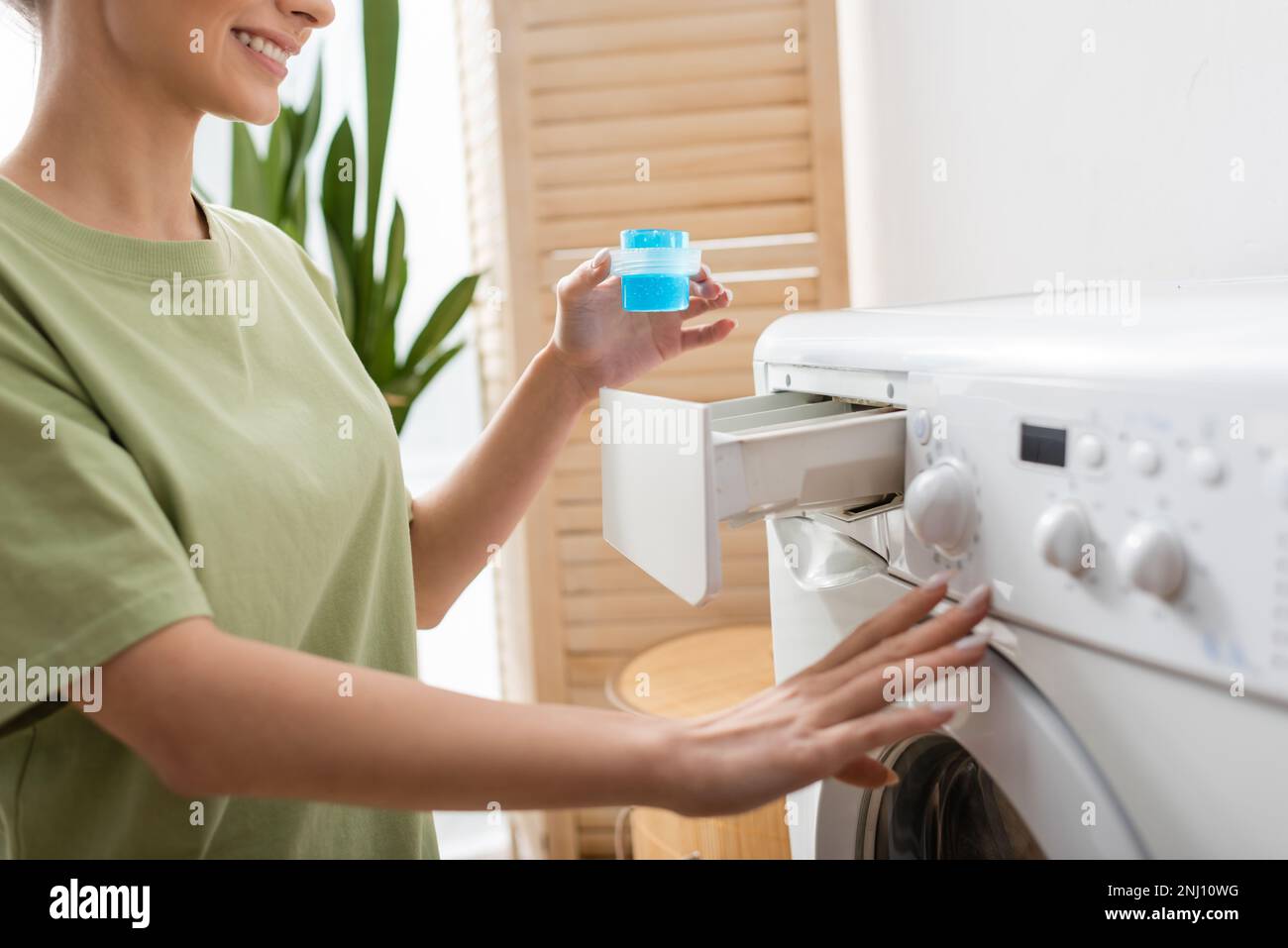 Vista ritagliata di donna sorridente che tiene liquido detergente vicino alla lavatrice a casa, immagine stock Foto Stock