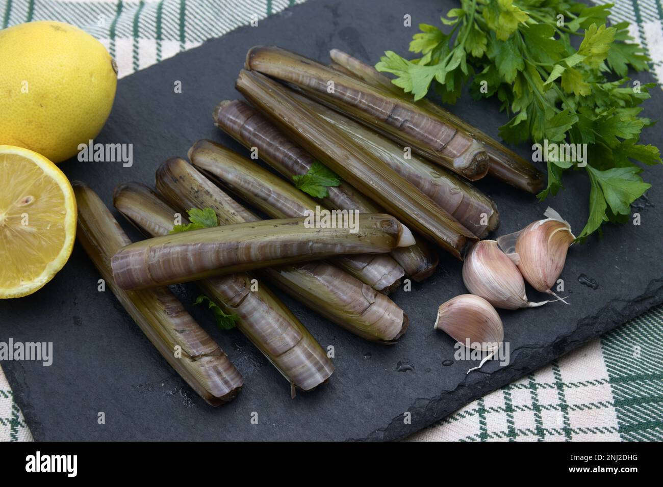 Vongole fresche di rasoio con aglio, prezzemolo e limone su piatto di ardesia nero. Presentazione perfetta per un'autentica esperienza di pesce. Foto Stock Vongole fresche di rasoio con aglio, prezzemolo e limone su piatto di ardesia nero. Presentazione perfetta per un'autentica esperienza di pesce. Foto Stock