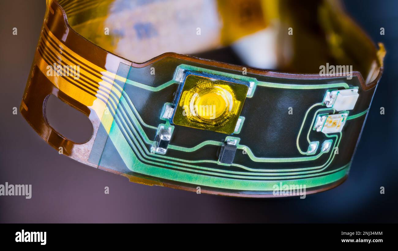 Circuito stampato flessibile e piccoli componenti elettronici in striscia di plastica ricurvi in cerchio su sfondo scuro. Primo piano del circuito stampato flessibile dalle cuffie. Foto Stock