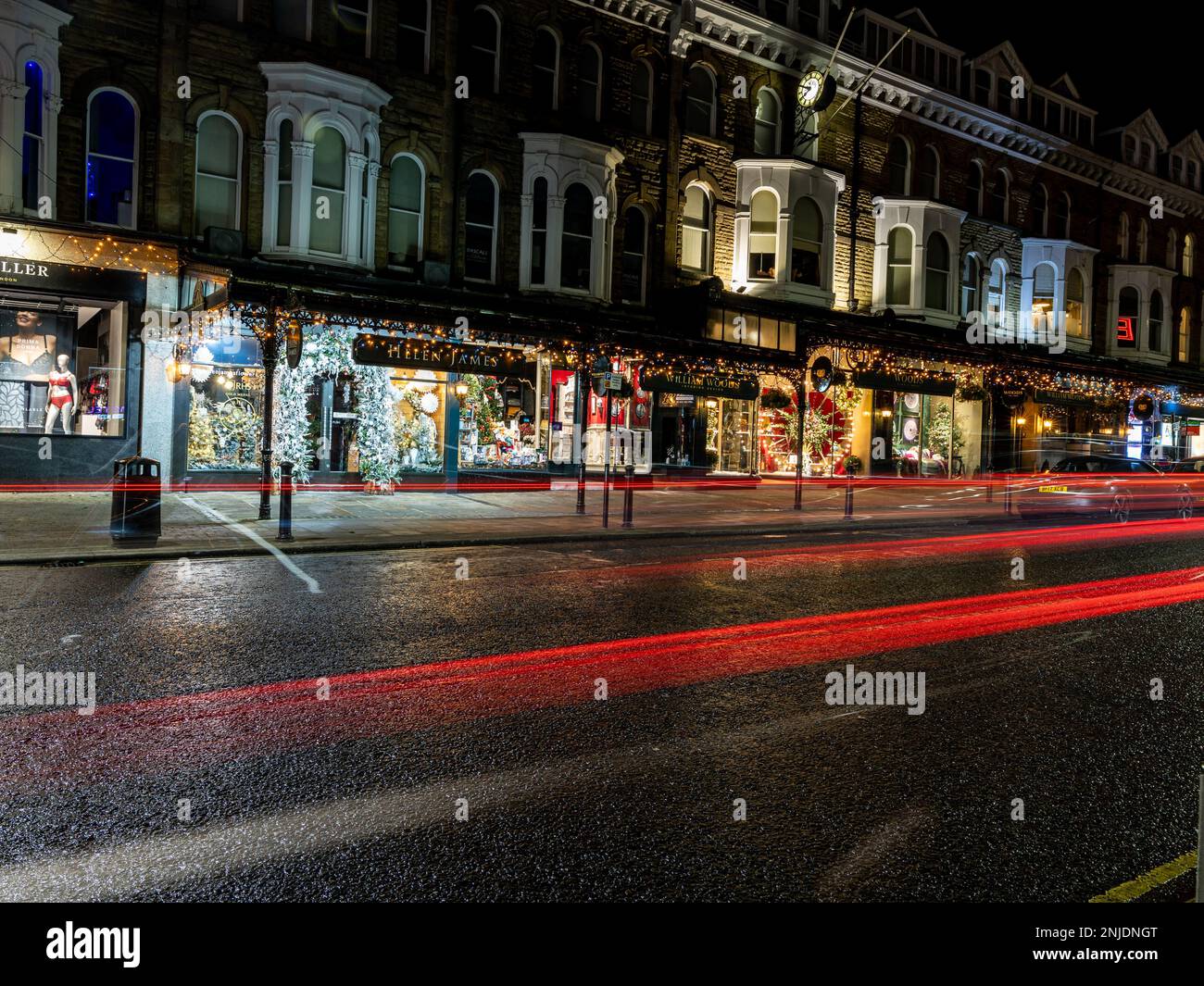 Vista notturna a lunga esposizione di una strada di Harrogate con tracce di luce rosse e bianche di auto, vetrine dei negozi illuminate e marciapiede bagnato, North Yorkshire Foto Stock