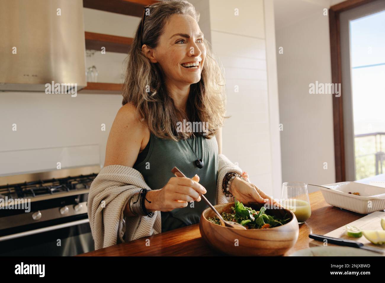 Donna matura sorridente felicemente mentre ha un pasto vegano sano nel paese. Donna anziana che si serve una sana ciotola di buddha nella sua cucina. Donna tak Foto Stock
