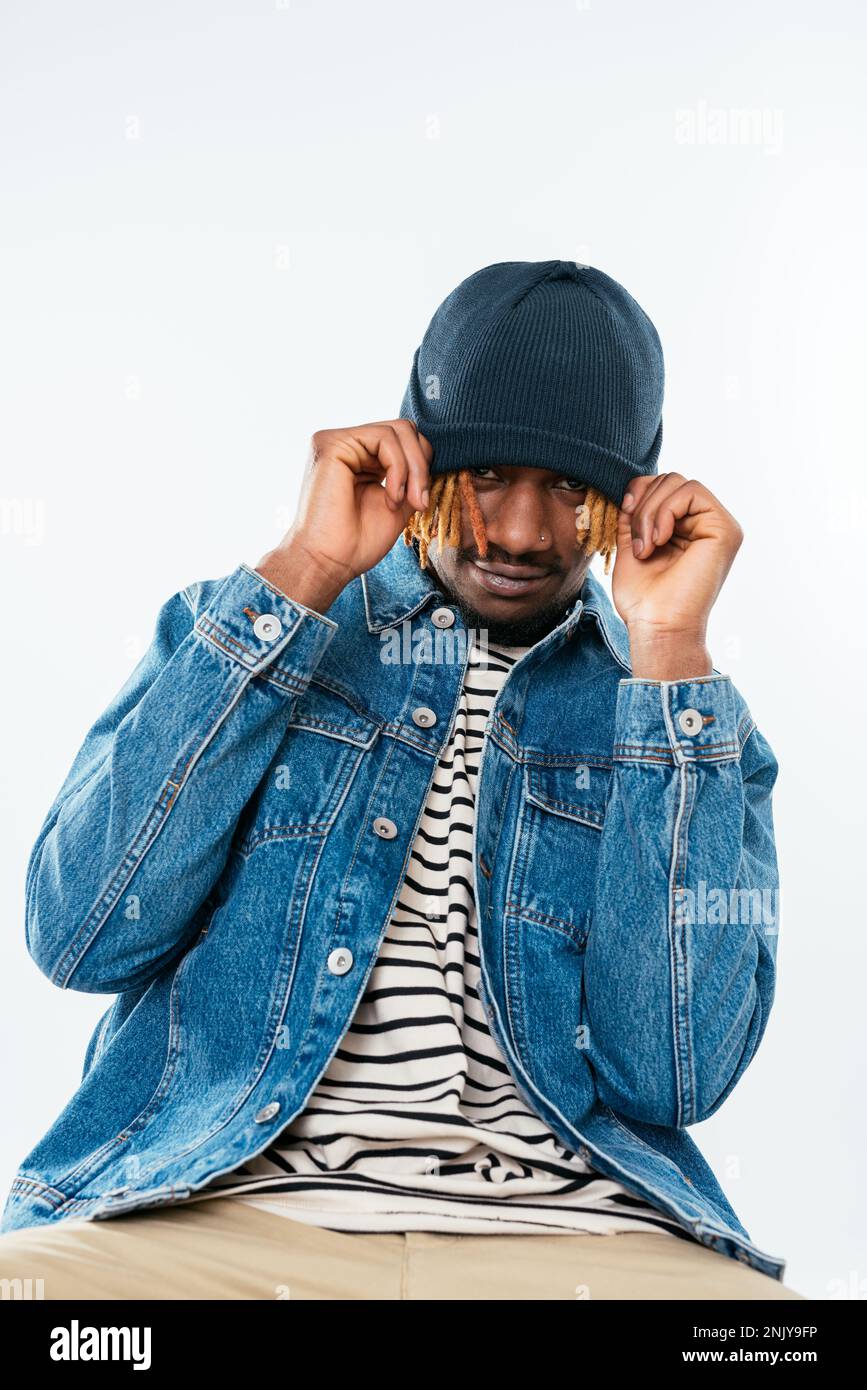 Giovane afro-americano positivo con baffi e lucchetti in giacca denim seduta su sedia e cappello a trazione sulla fronte in studio Foto Stock