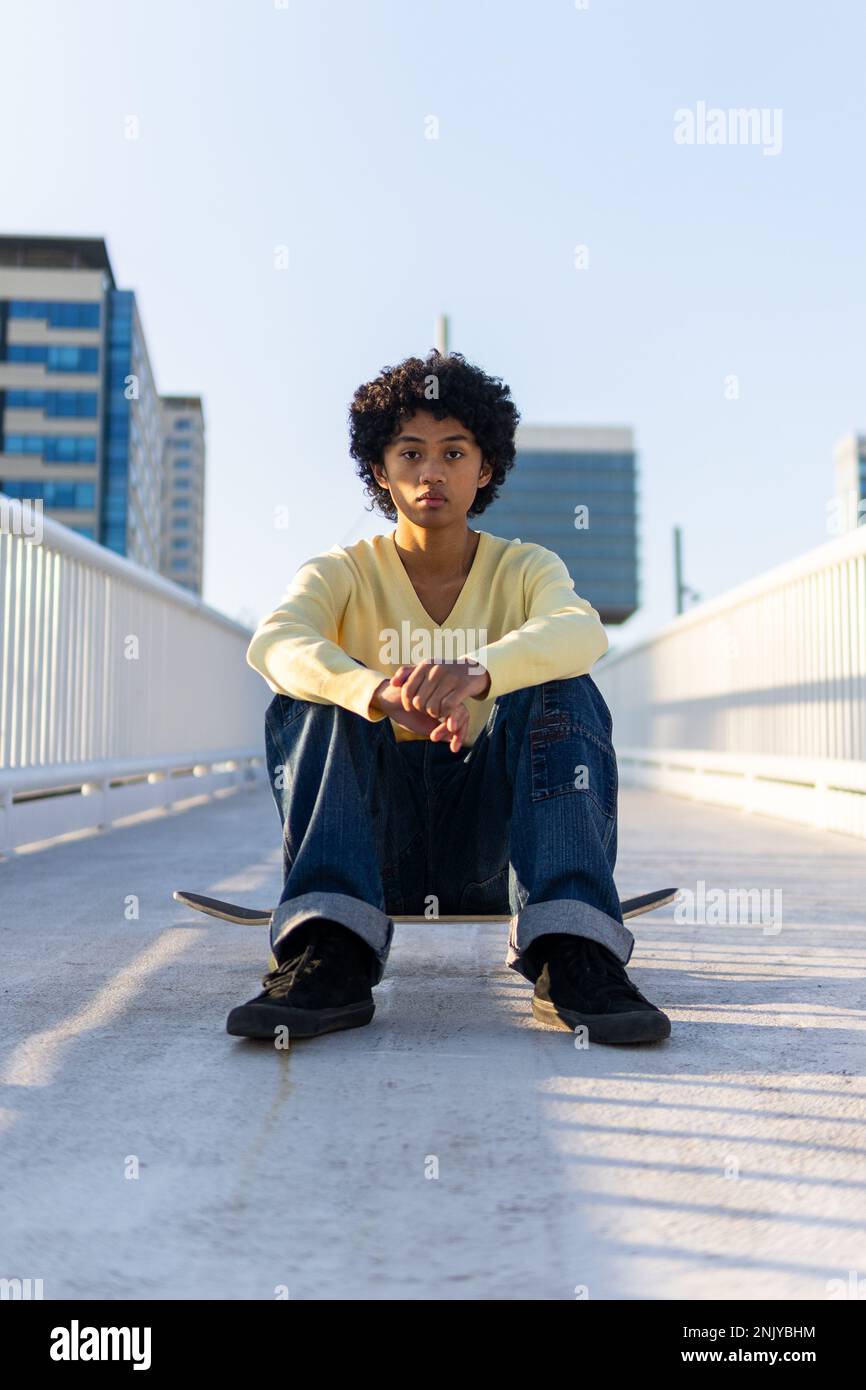 Corpo pieno di serio giovane skater asiatico maschio in abito casual guardando la macchina fotografica mentre si siede sullo skateboard sul ponte Foto Stock