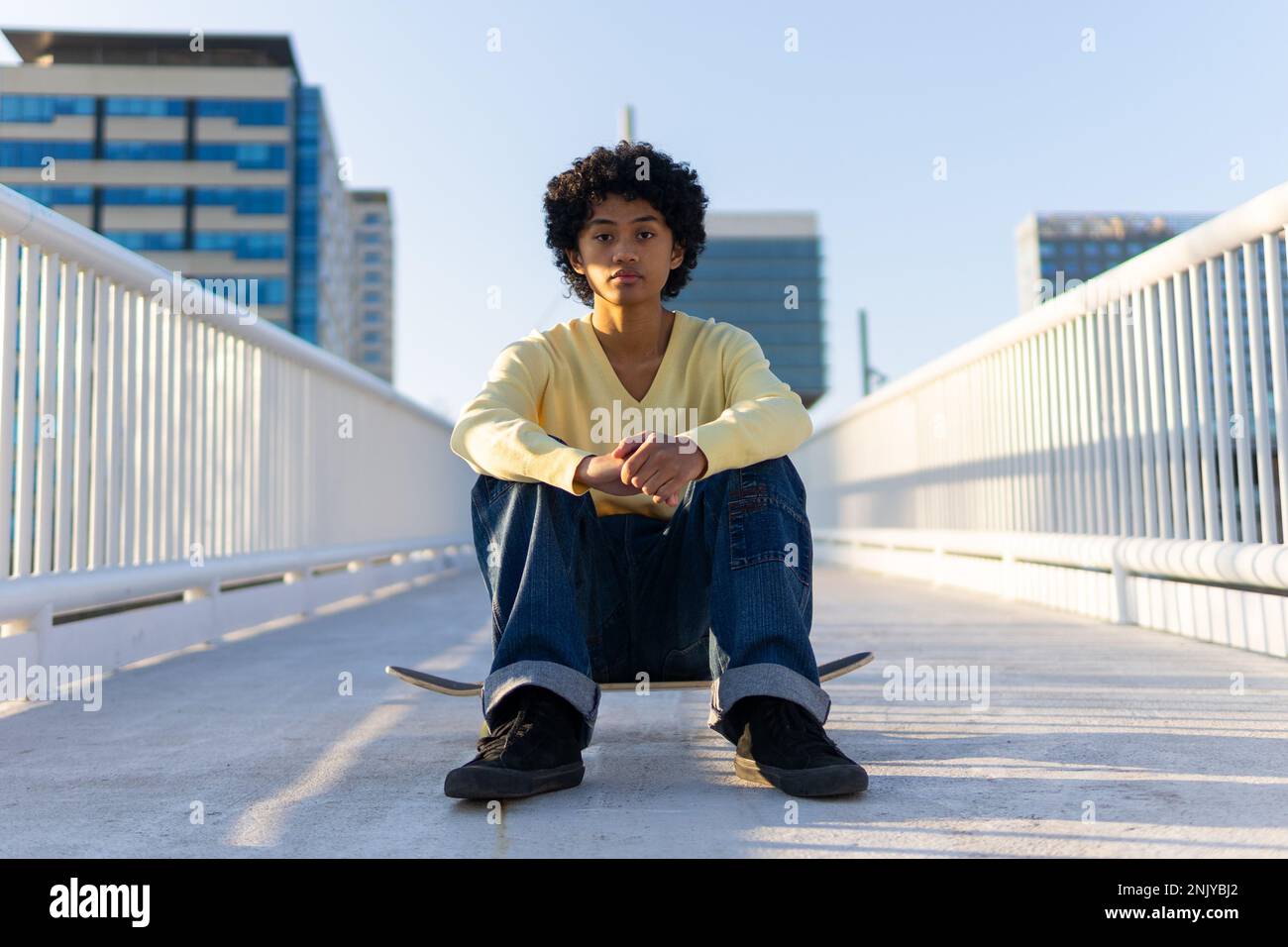 Corpo pieno di serio giovane skater asiatico maschio in abito casual guardando la macchina fotografica mentre si siede sullo skateboard sul ponte Foto Stock