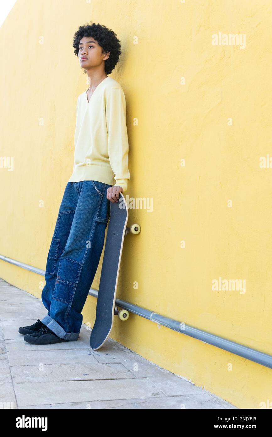Corpo pieno di giovane skater asiatico in abiti casual con acconciatura afro in piedi vicino a una parete gialla luminosa con skateboard e look awa Foto Stock