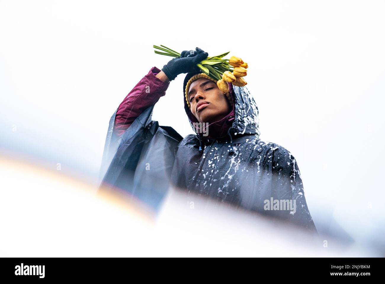 Basso angolo di giovane afro-americana femmina in impermeabile con gli occhi chiusi e tenendo tulipani gialli contro il cielo nuvoloso Foto Stock