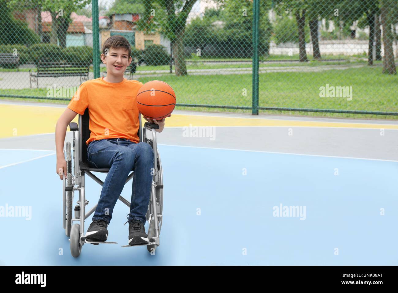 Ragazzo minorato in sedia a rotelle con palla da basket nel campo all'aperto Foto Stock