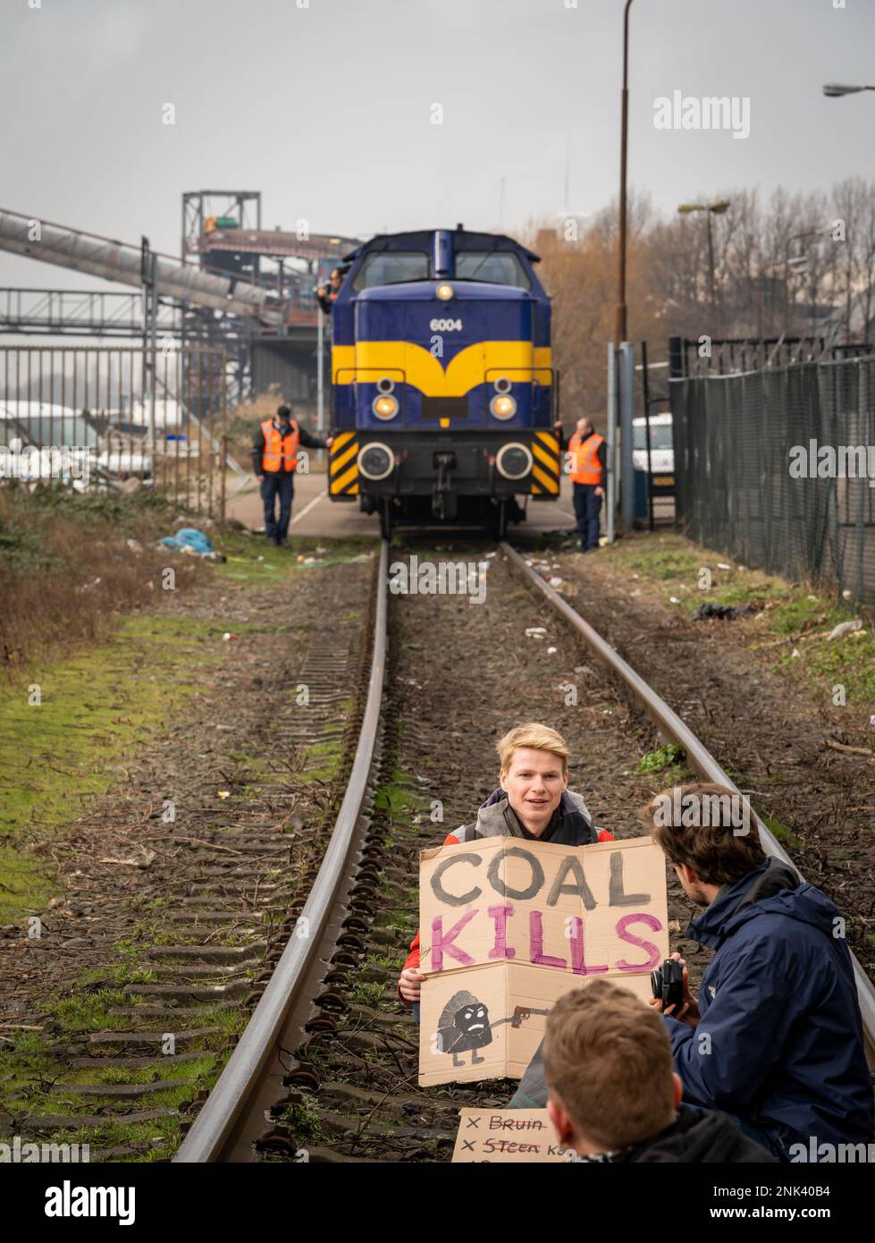 Amsterdam, Paesi Bassi, 22.02.2023, giovane attivista del clima con un cartello che dice "Coal Kills” seduto su una ferrovia di fronte al treno del carbone Foto Stock