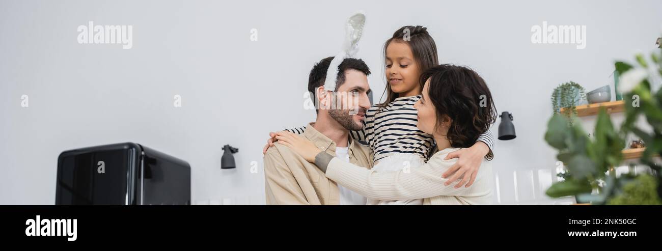 Donna sorridente e figlia che abbraccia l'uomo con la fascia di Pasqua in cucina, banner Foto Stock