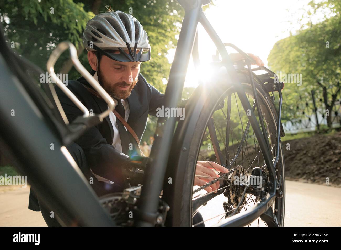 Giovane uomo d'affari bearded che controlla la bicicletta prima di andare a cavallo per lavorare la mattina. Vista frontale di un bel dipendente maschio nel casco che ispeziona la ruota della bicicletta, WHI Foto Stock