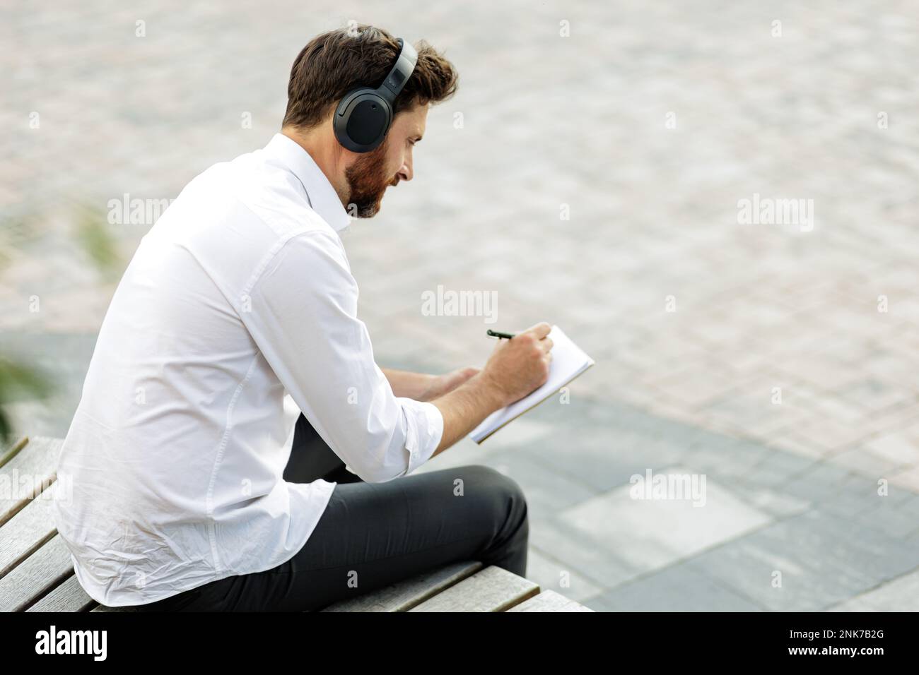 Uomo elegante concentrato in cuffie wireless scrittura nuovo business plan in blocco note. Direttore dai capelli scuri con musica d'ascolto setola mentre si siede sul woo Foto Stock