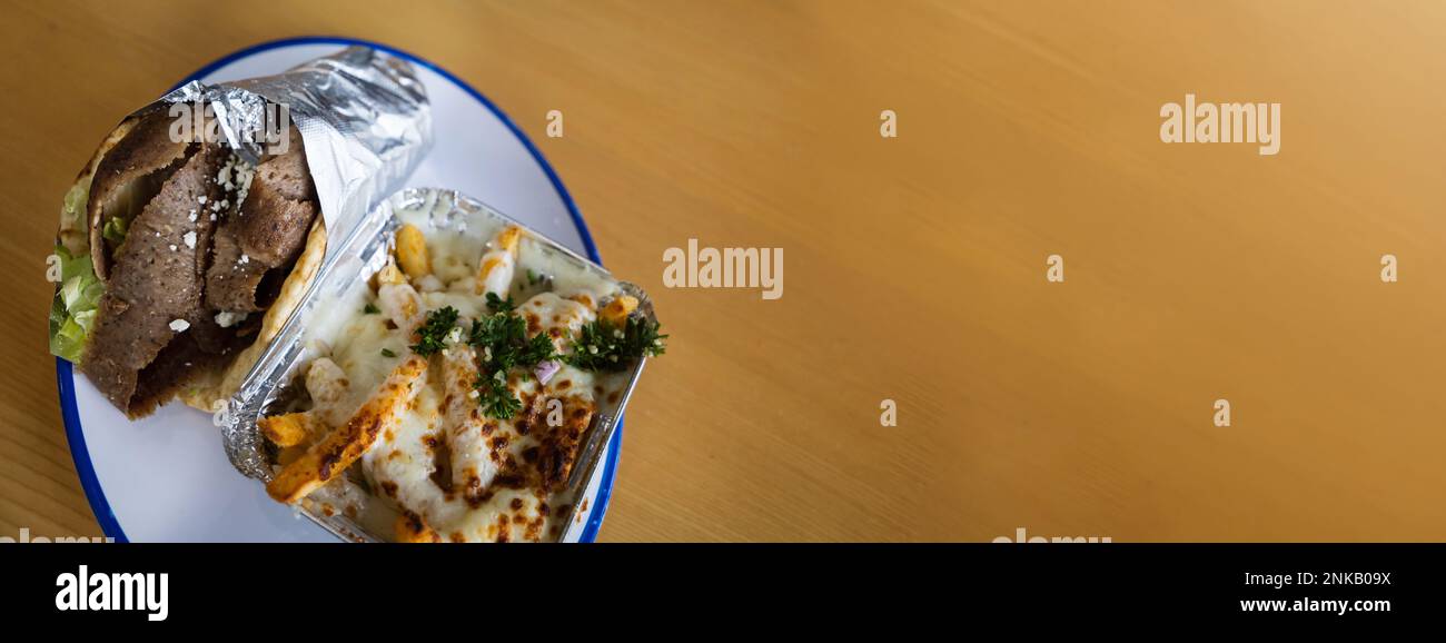 Un giroscopio greco mediterraneo avvolto in un foglio su un piatto con un lato di patatine fritte greche ricoperte di mozzarella e formaggio feta fuso, erbe aromatiche e spezie Foto Stock