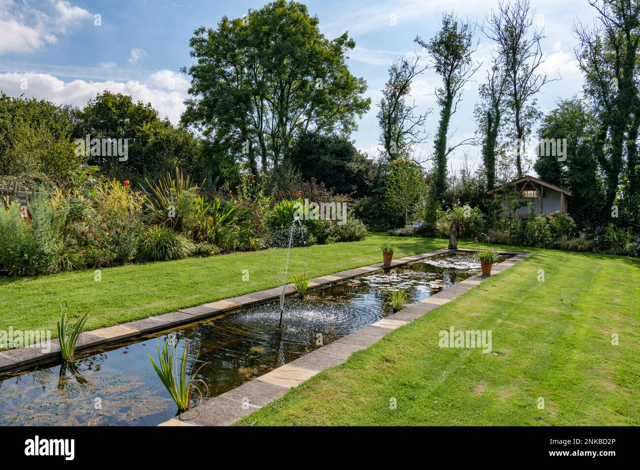 Scena estiva guardando attraverso un prato con un rill in un giardino di campagna inglese con una piccola fontana che gioca e delicati riflessi Foto Stock