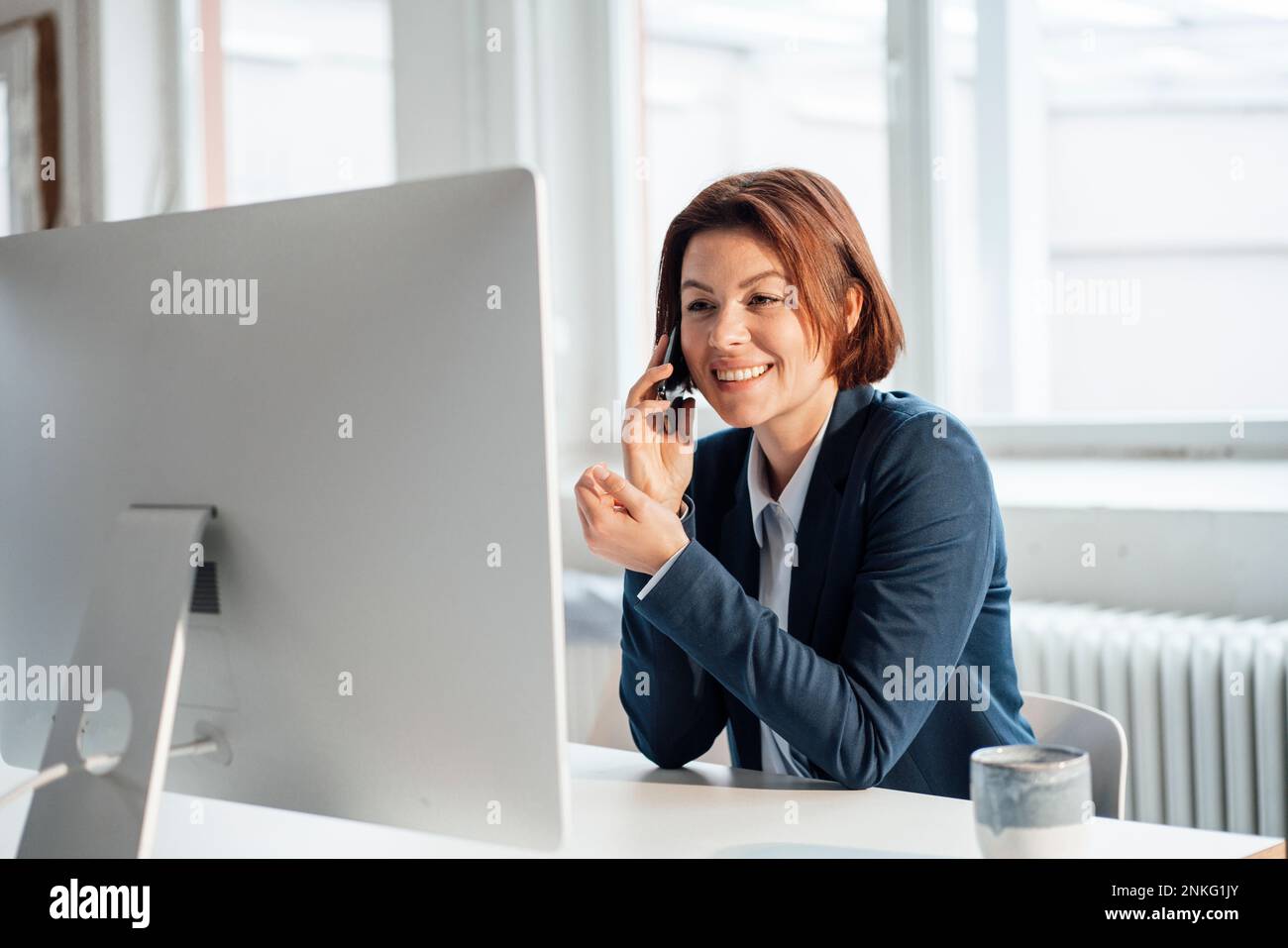 Donna d'affari felice che parla sullo smartphone davanti al desktop sul posto di lavoro Foto Stock