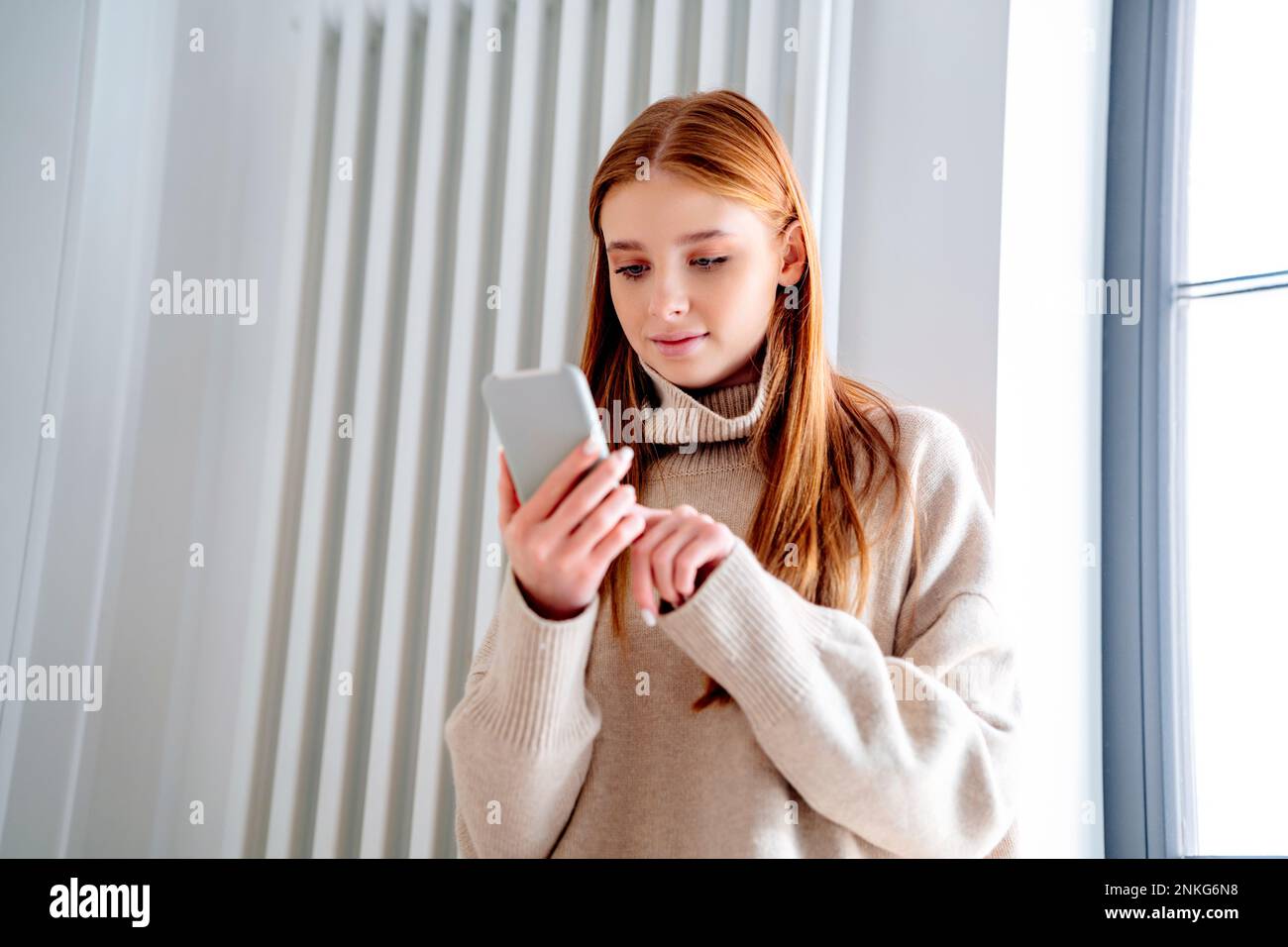 Ragazza giovane sorridente che usa lo smartphone di fronte al radiatore a casa Foto Stock