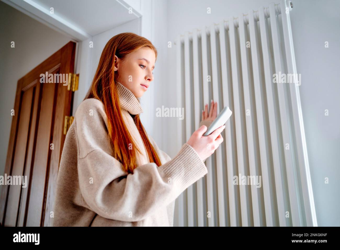Ragazza adolescente che usa il telefono cellulare per controllare la temperatura del radiatore a casa Foto Stock