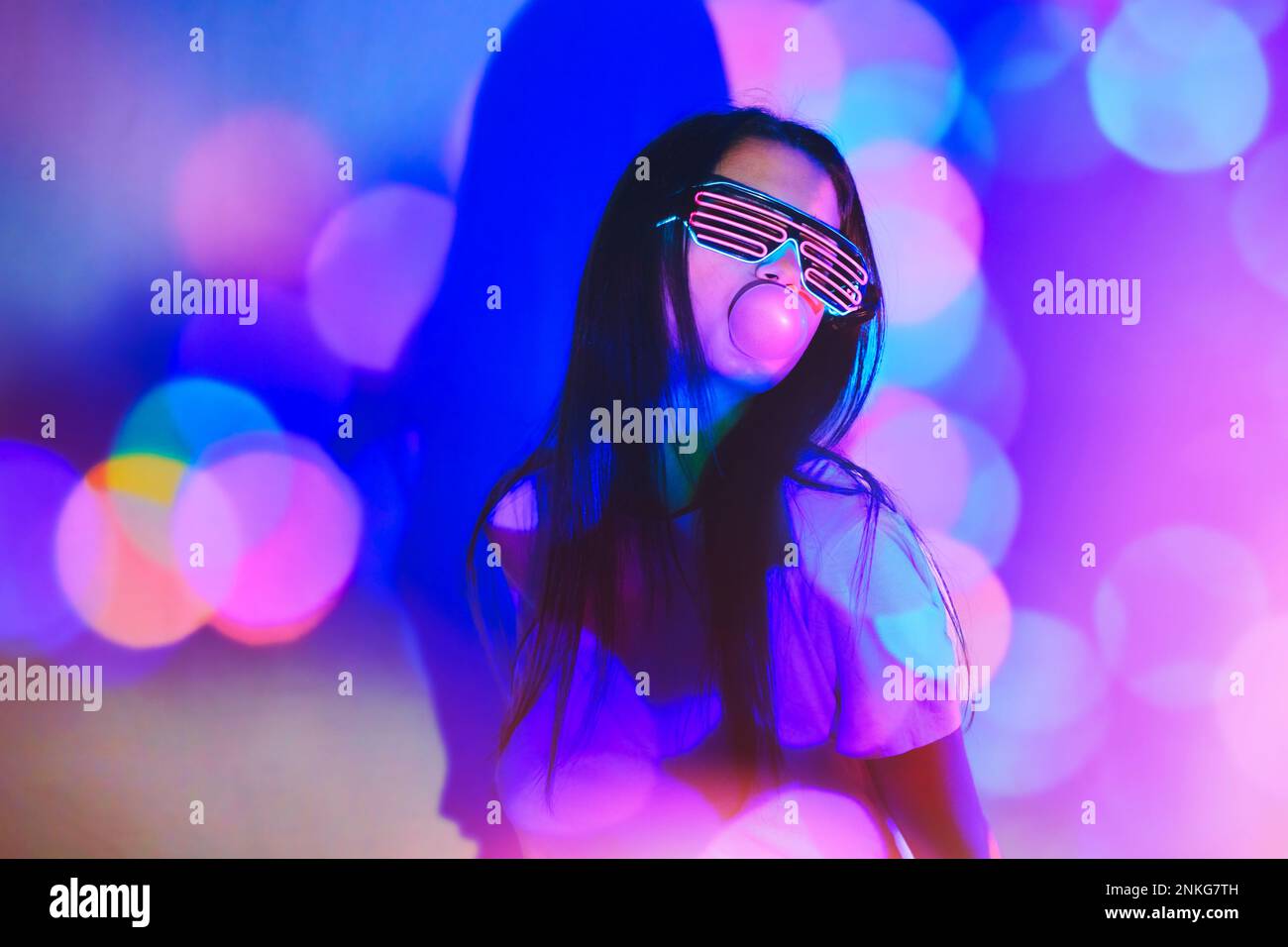 Giovane donna con occhiali futuristici che soffiano la bolla su sfondo colorato al neon Foto Stock