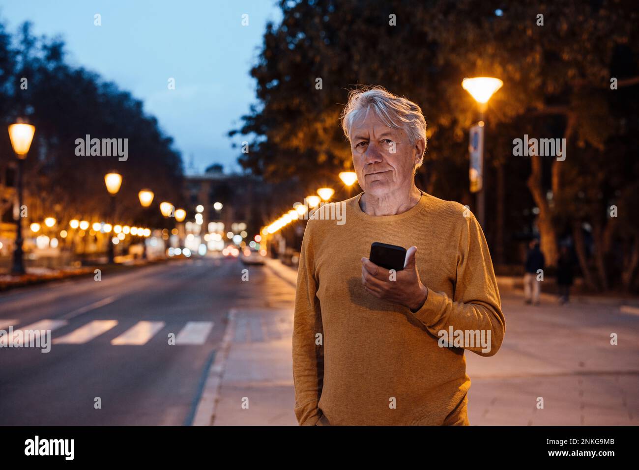 Uomo anziano sorridente in piedi con il telefono cellulare sul marciapiede Foto Stock
