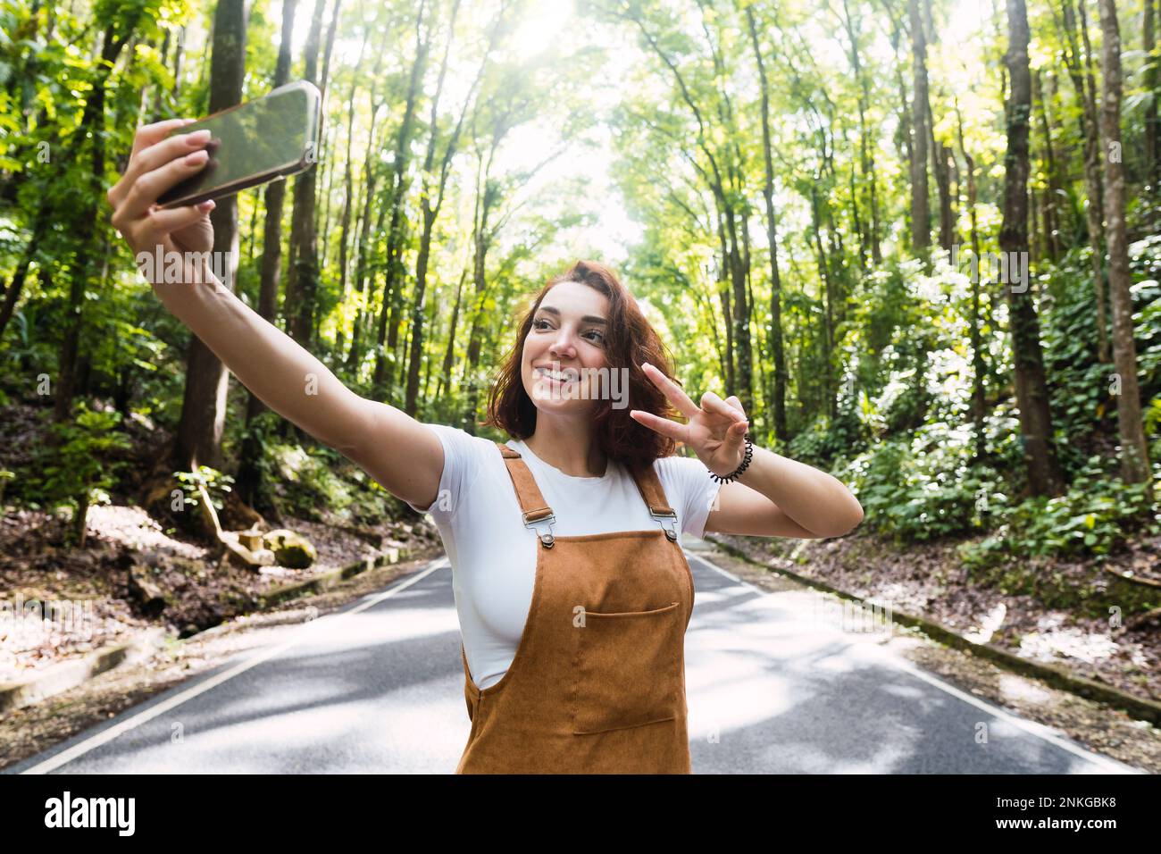 Felice giovane donna che mostra segno di pace gesto e di presa selfie di fronte agli alberi Foto Stock