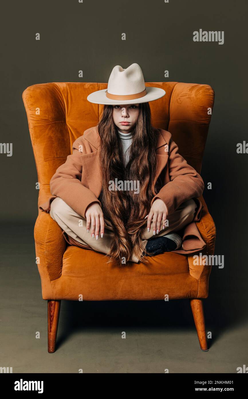 Ragazza adolescente che indossa cappello seduto su poltrona contro sfondo grigio Foto Stock