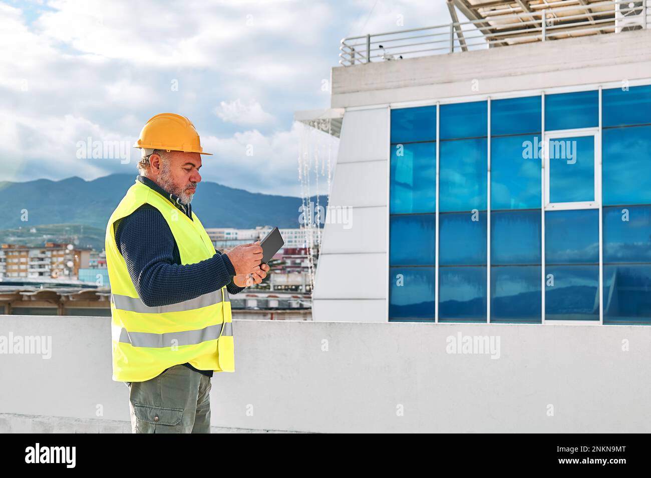 Supervisore al ribasso di mezza età in elmetto e giubbotto di sicurezza con tablet sul sito dell'edificio. L'ingegnere strutturale o architetto monitora l'avanzamento di Foto Stock