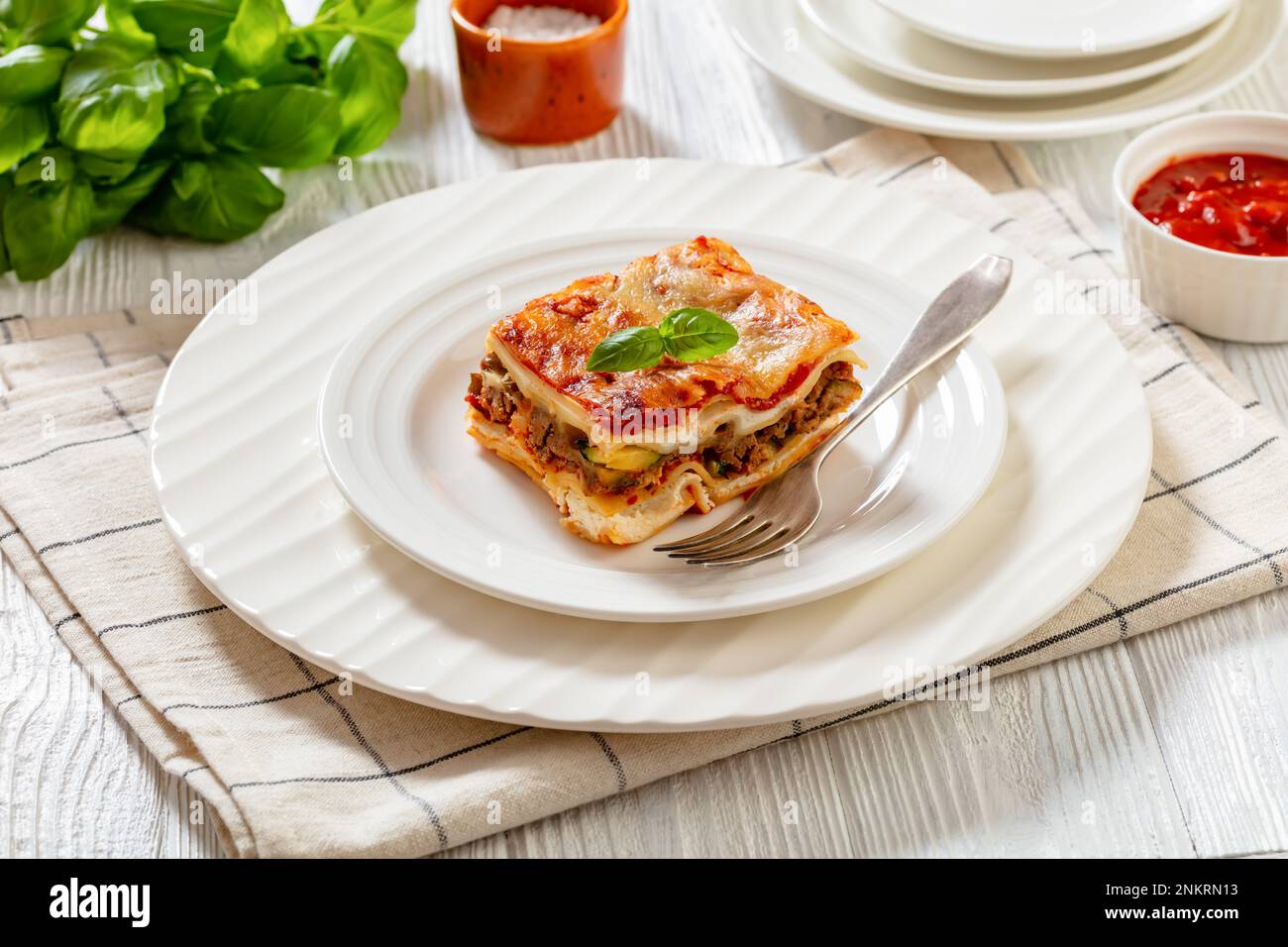 Porzione di lasagna ricoperta di ricotta, carne macinata, funghi, zucchine e salsa di pomodoro su piatto bianco con forchetta su tavola di legno bianco, terra Foto Stock