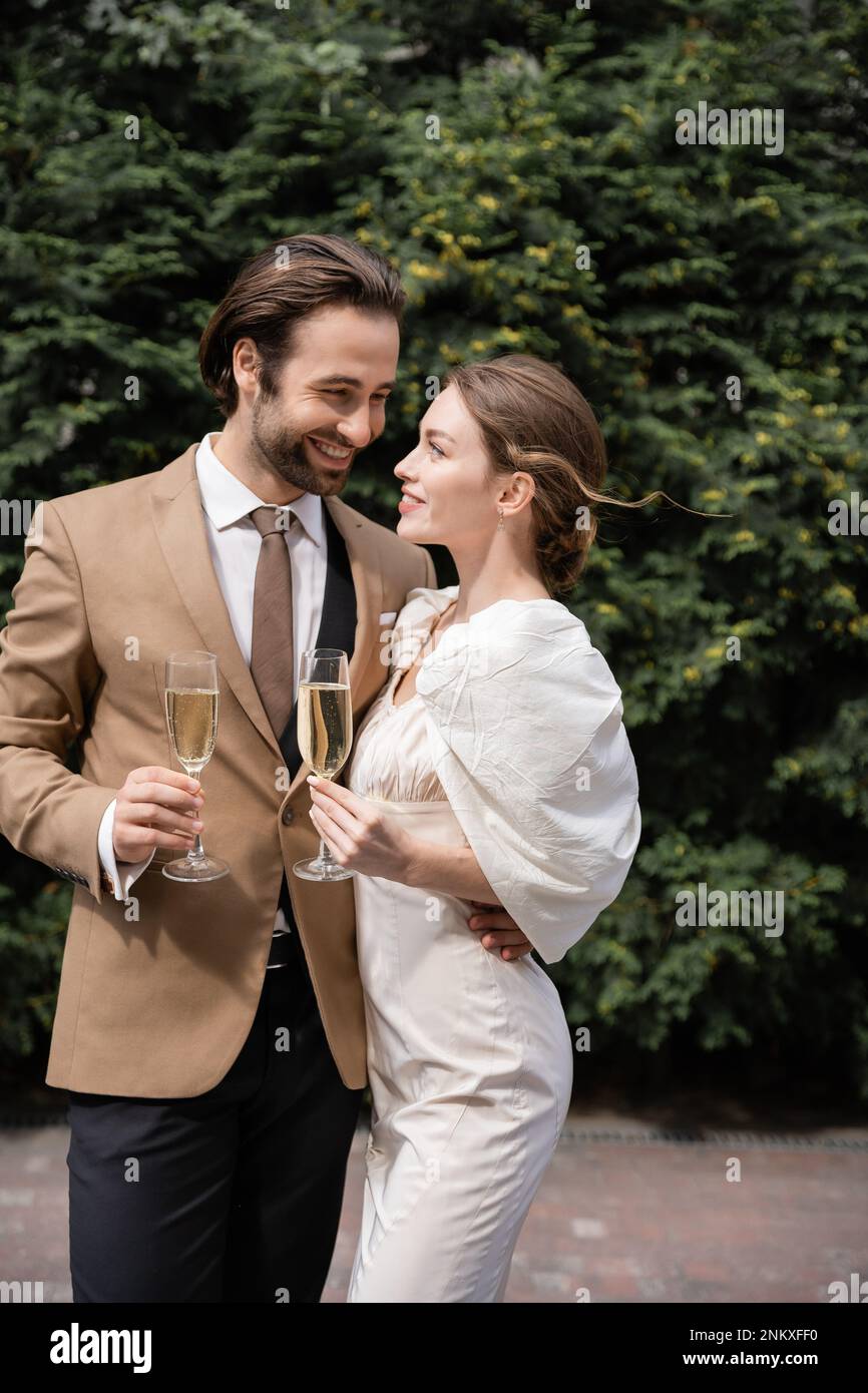 sposo allegro in tuta e sposa felice in abito da sposa con bicchieri di  champagne, immagine stock Foto stock - Alamy