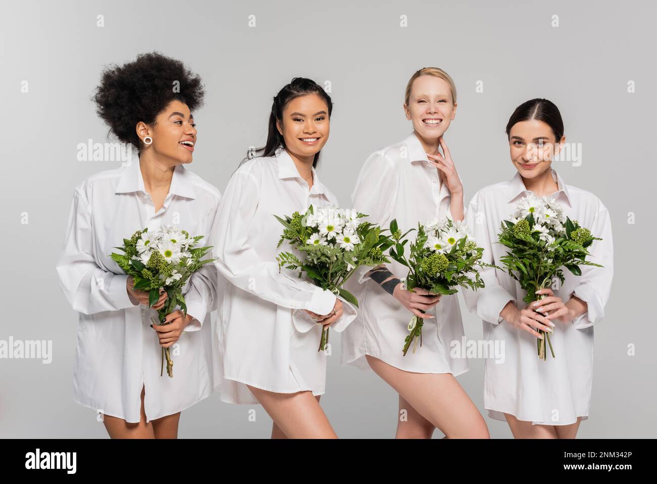 donne felice multiculturale in camicie bianche che tengono bouquet primavera isolato su grigio, immagine stock Foto Stock