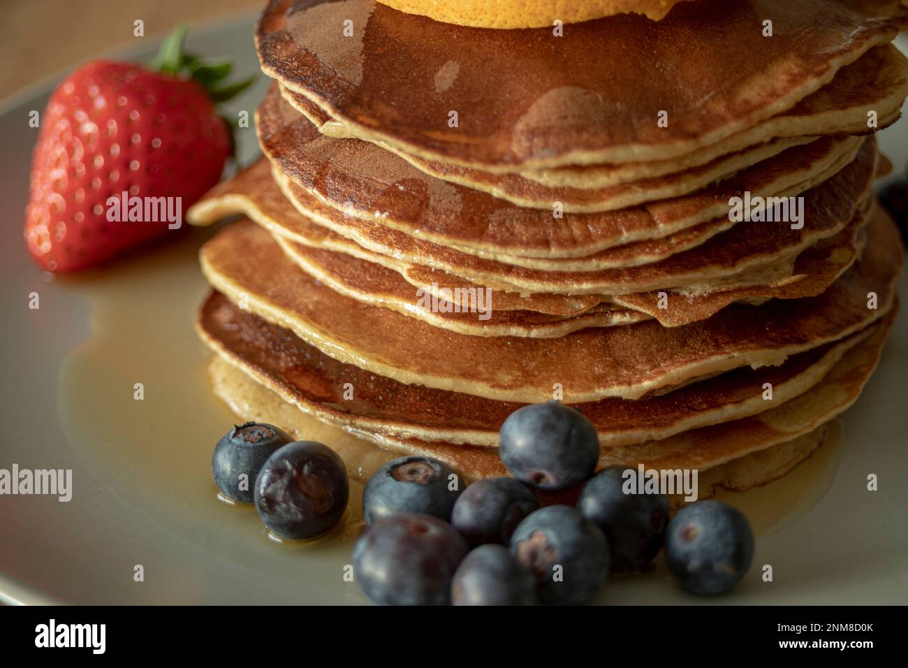 Primo piano di deliziosi pancake su un piatto con sciroppo, mirtilli, una fragola e una fetta d'arancia in un ambiente ben illuminato Foto Stock