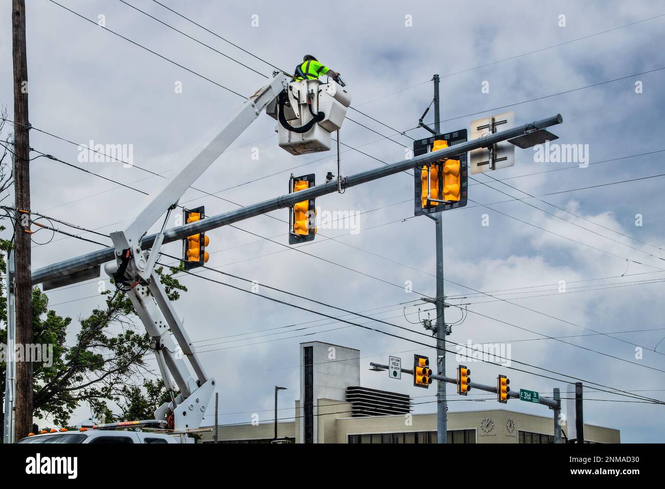 Operaio in alto nella benna della gru che ripara il semaforo all'incrocio della città nelle giornate nuvolose con l'edificio in background Foto Stock