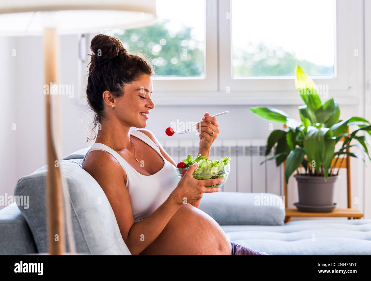 Bella giovane donna che mangia insalata sul divano a casa. Stile di vita sano durante il periodo di maternità Foto Stock