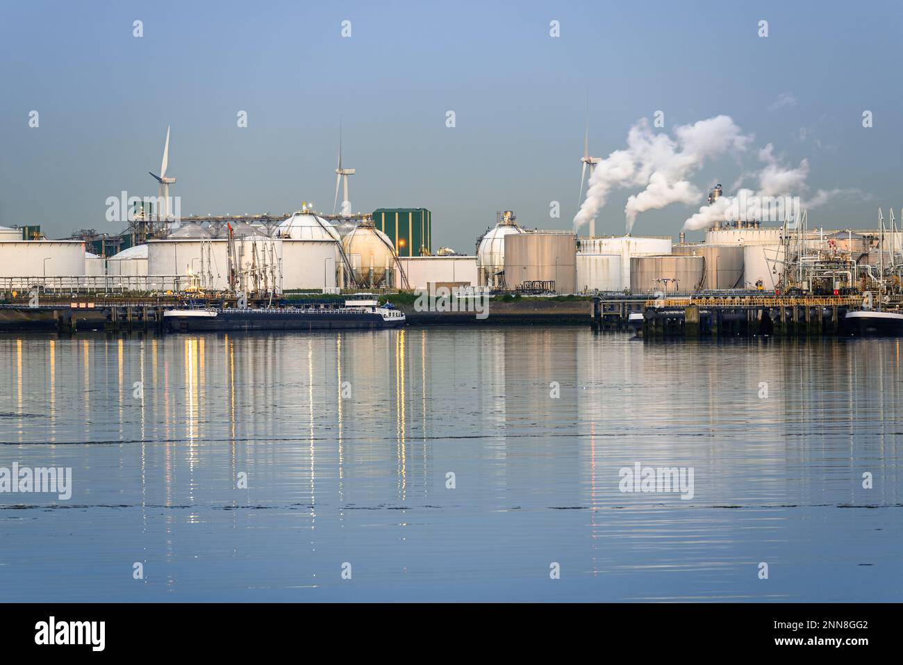 Raffineria di petrolio Harbourside con ciminiere di fumo bianco al crepuscolo. Le turbine eoliche sono in background. Foto Stock