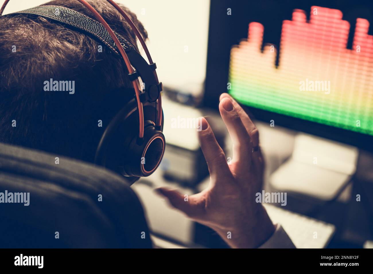 Vista posteriore di Electronic Music Producer seduto di fronte al suo computer in cuffie ascolto di brani appena creati. Foto Stock