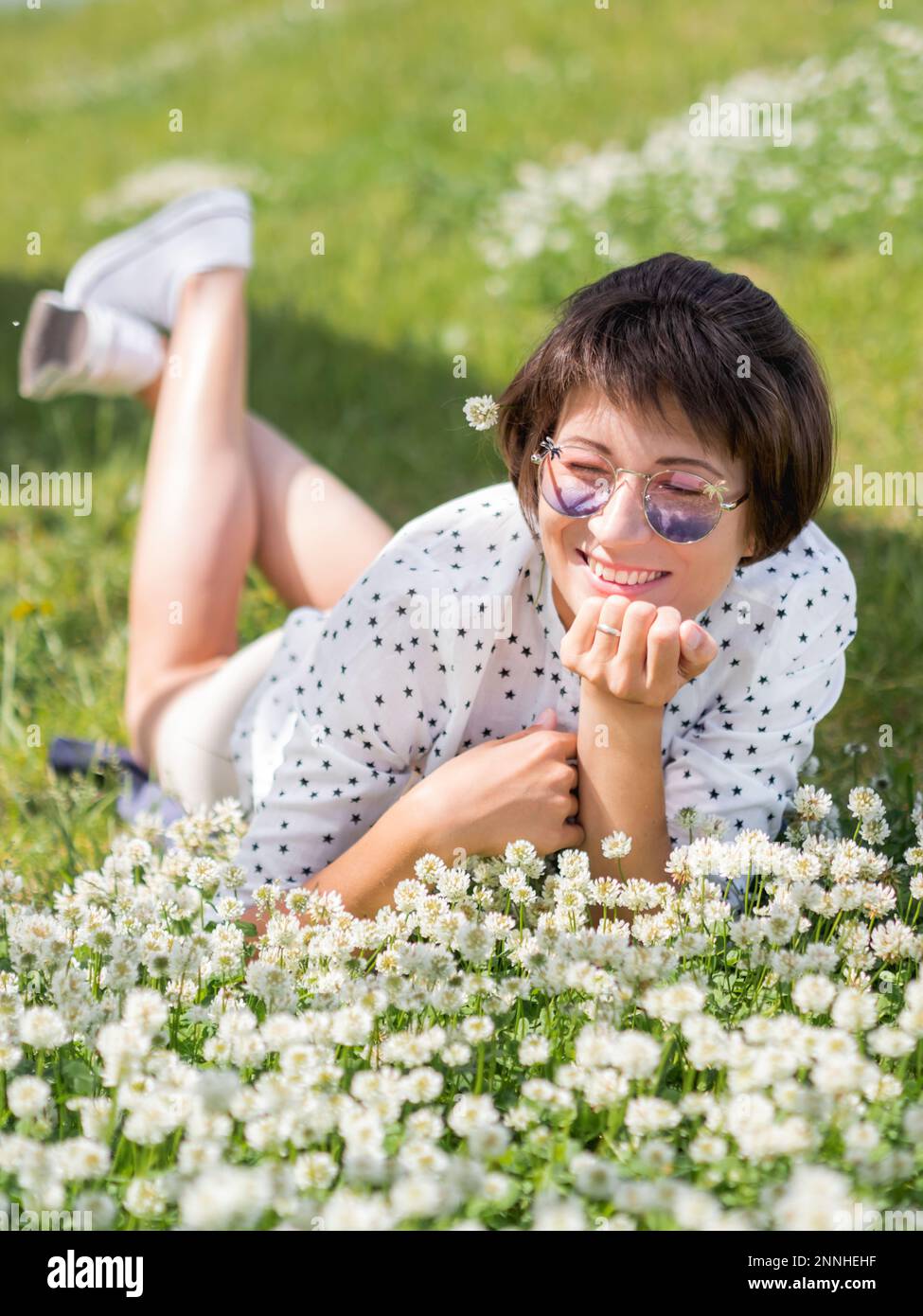 Donna in occhiali da sole colorati sniffs fiori trifoglio sul prato in parco urbano. Natura in città. Rilassati all'aperto dopo il lavoro. Atmosfera estiva. Foto Stock