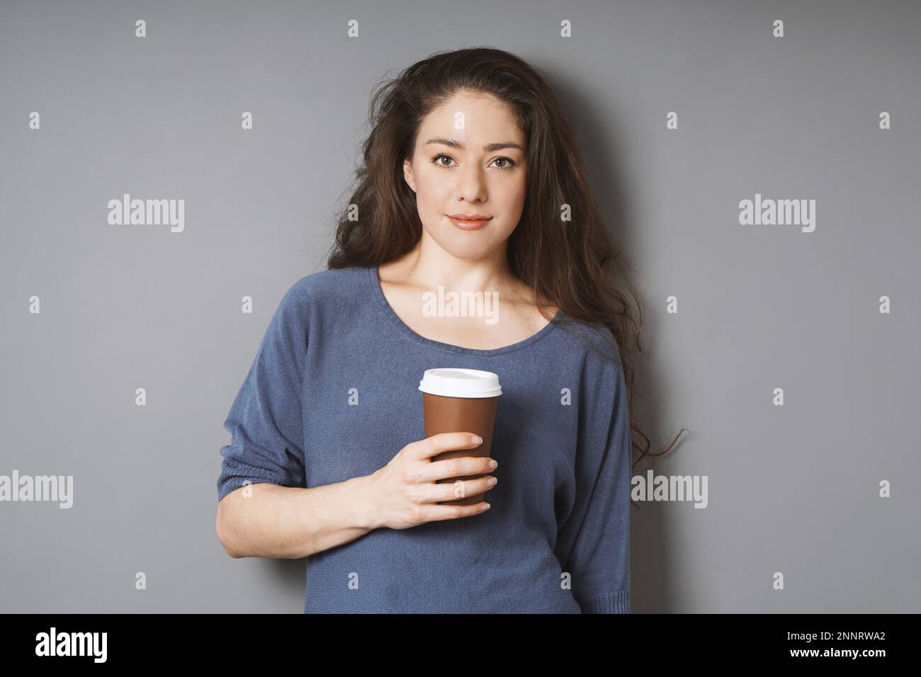 Rilassata giovane donna sulla sua pausa caffè - caffè di contenimento per andare in carta monouso bicchiere mentre appoggiata contro il muro grigio Foto Stock
