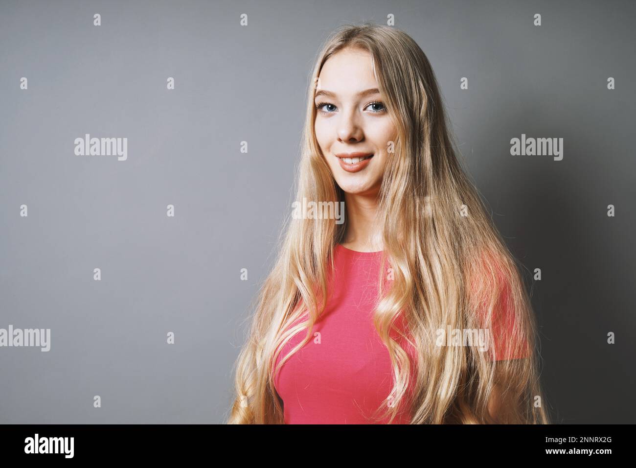 Sorridenti giovane donna con capelli lunghi biondi e t-shirt rosa contro uno sfondo grigio con spazio di copia Foto Stock