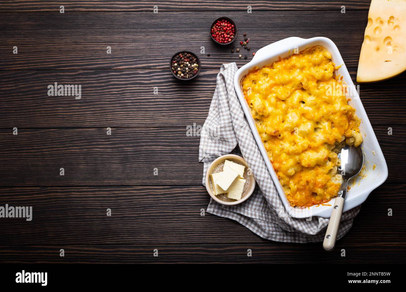 Primo piano di maccheroni al forno e formaggio in casseruola bianca su fondo ligneo, con condimenti, burro, cucchiaio e forchetta, vista dall'alto, spazio per il testo. Foto Stock