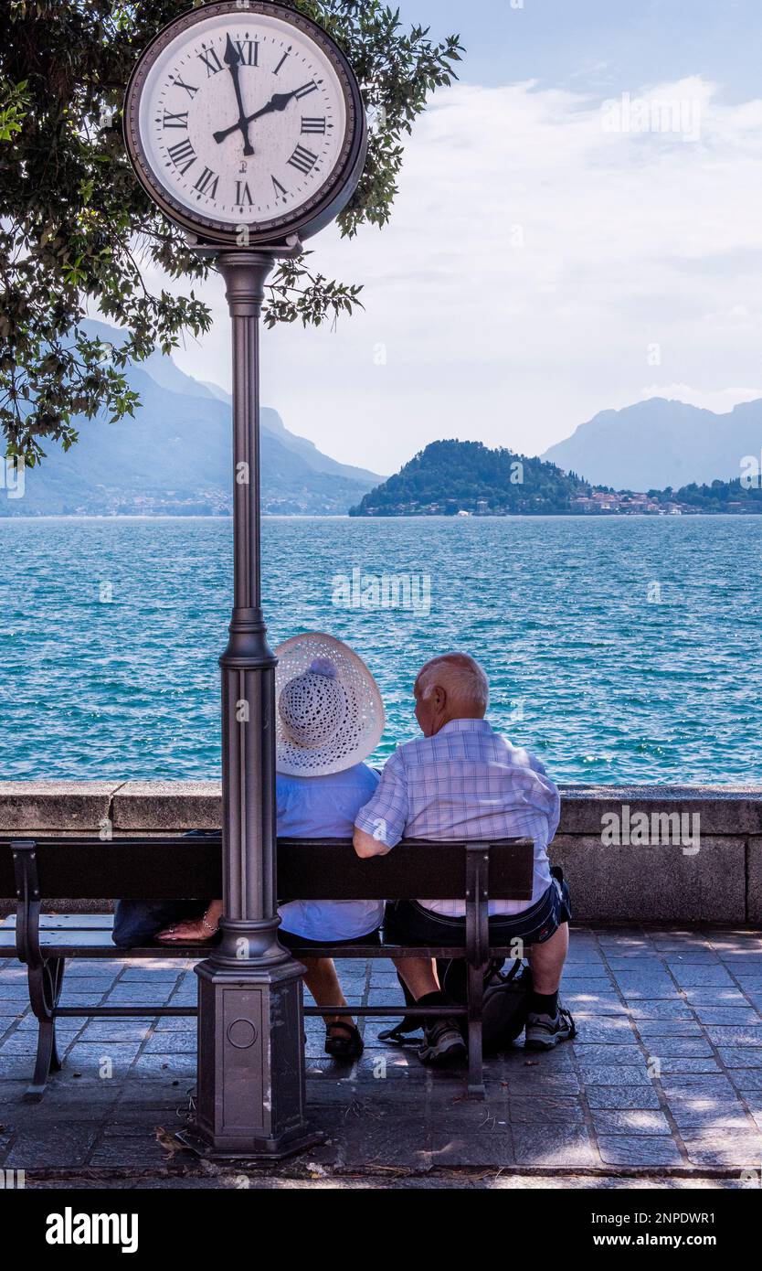 Una vecchia coppia guarda il Lago di Como da una panchina di Menaggio con un orologio di strada proprio dietro di loro. Foto Stock