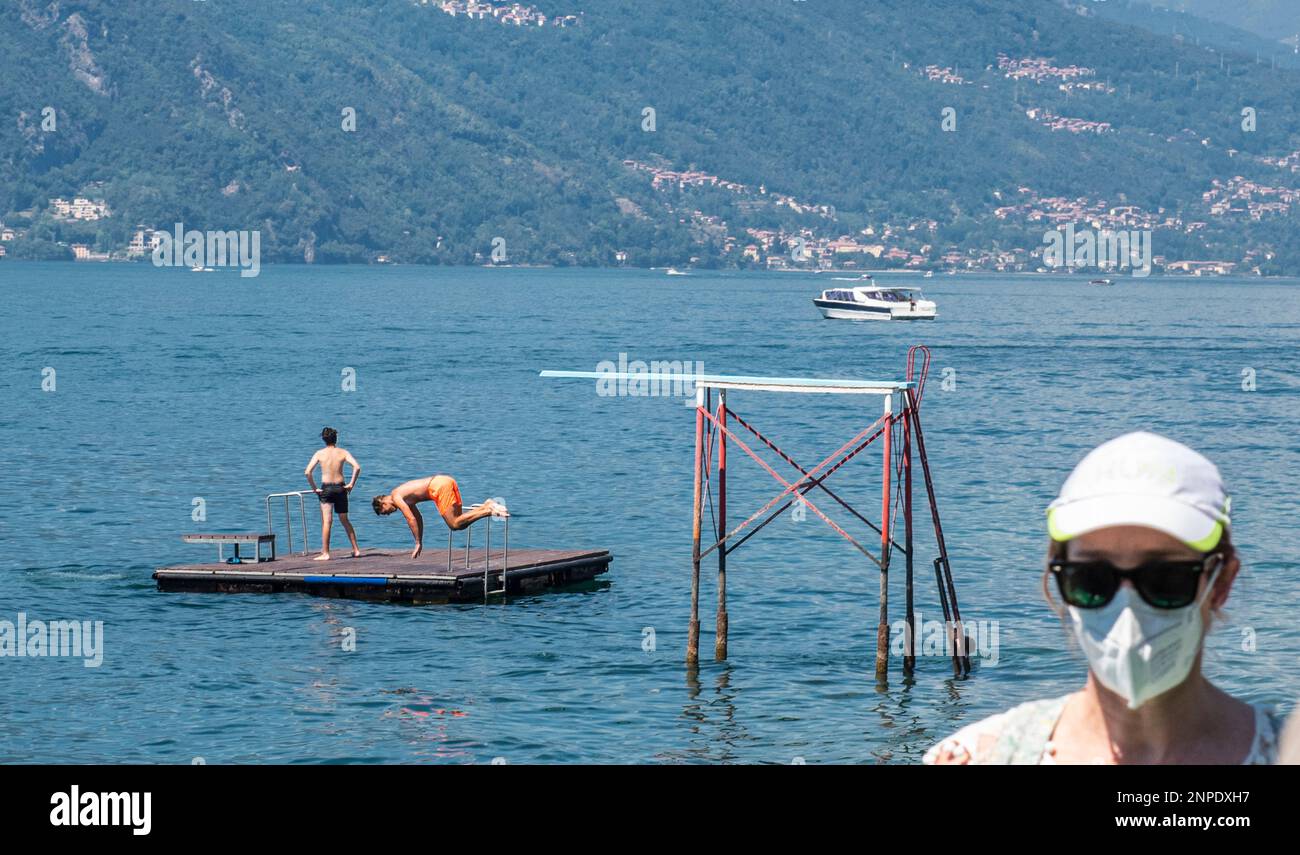 Un uomo viene catturato a mezz'aria mentre salta in acqua sul lago di Como mentre una donna che indossa occhiali da sole e una maschera facciale entra nella cornice in primo piano. Foto Stock